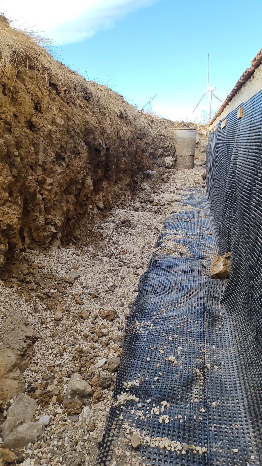 Trincea con ghiaia, membrana impermeabile nera e terra contro un cielo azzurro.