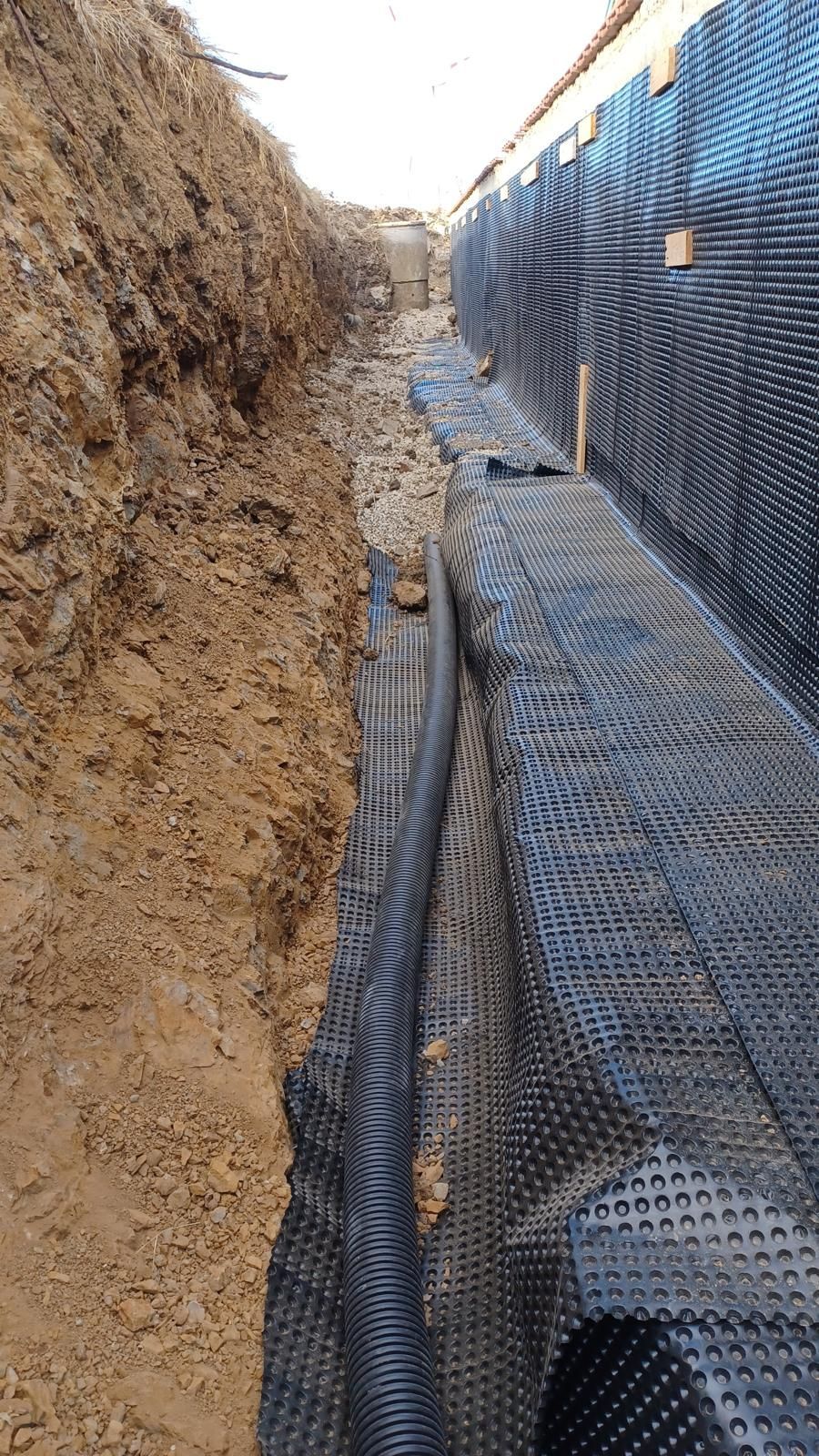 Una trincea che mostra l'impermeabilizzazione delle fondamenta con membrana di drenaggio bugnata contro un muro e riempita con ghiaia.
