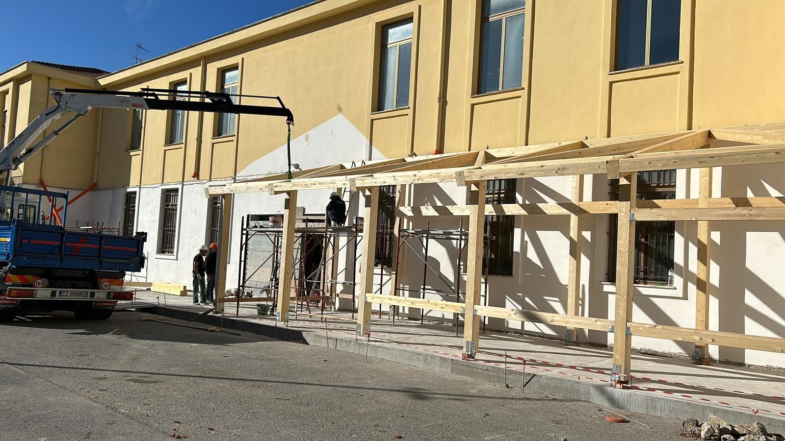 Lavori in corso: impalcature, camion, edificio con pareti gialle e bianche, struttura in legno per un pergolato.