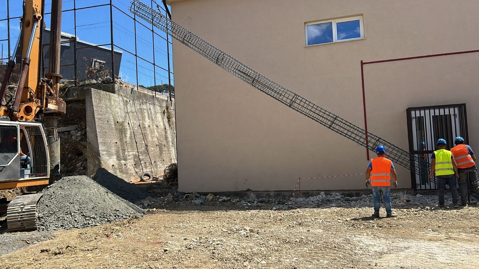 Cantiere edile con macchinari pesanti, operai con giubbotti antiproiettile e un edificio in costruzione.