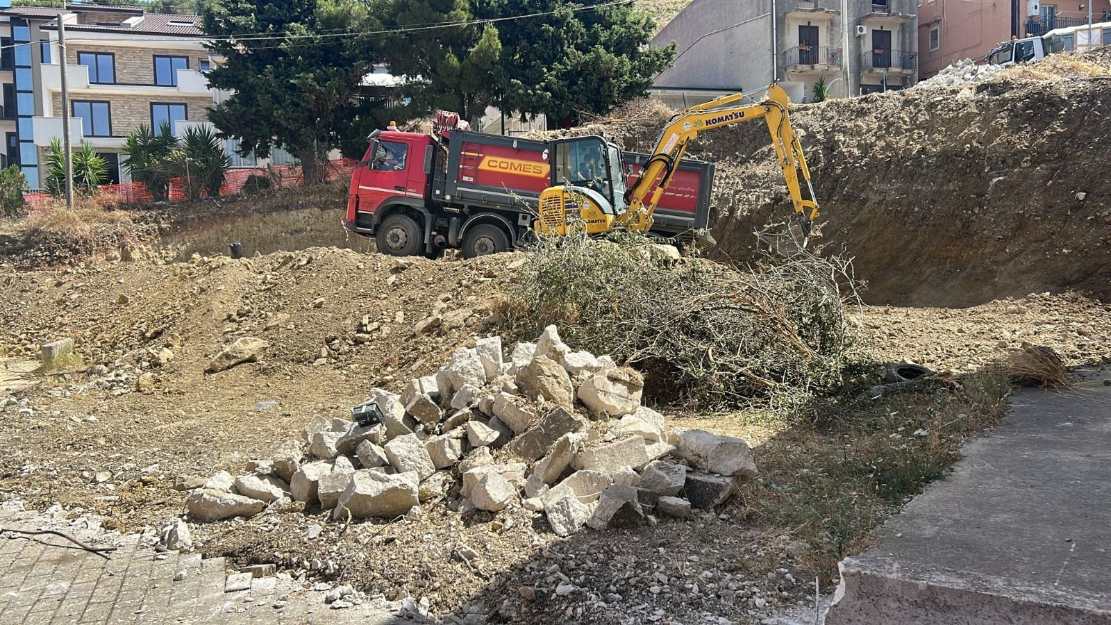Un escavatore carica un autocarro con cassone ribaltabile pieno di terra e detriti in un cantiere edile.