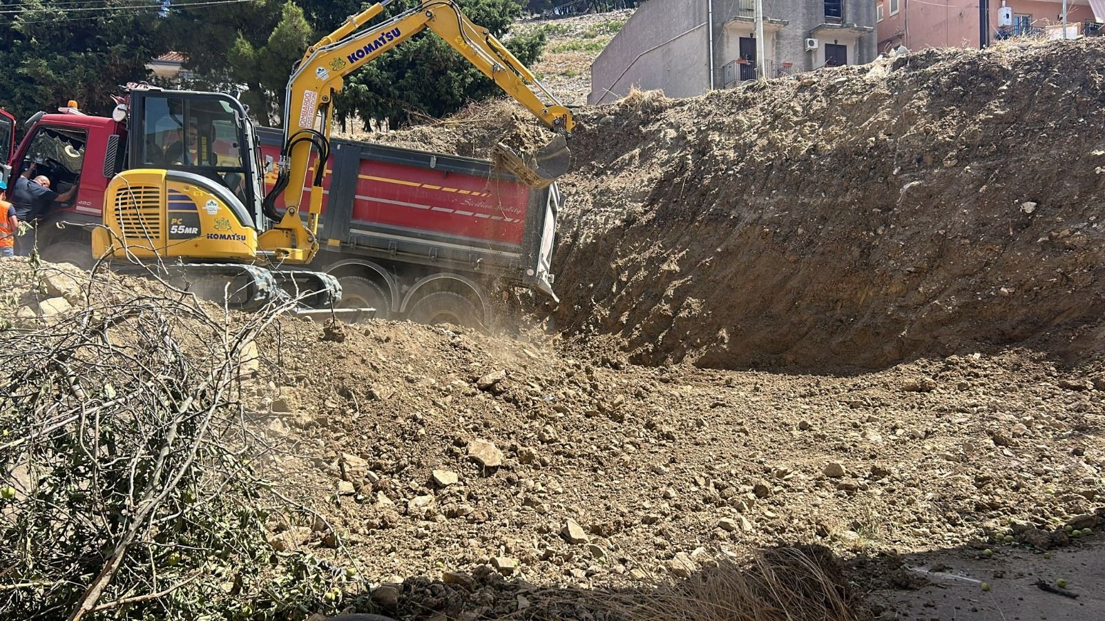 Un escavatore giallo carica un camion di macerie; giorno, all'esterno.