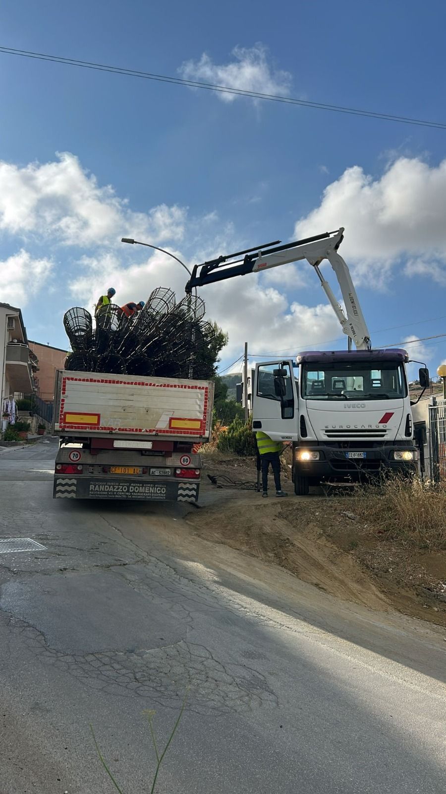 Camion che carica detriti con un braccio meccanico su una strada sotto un cielo azzurro.