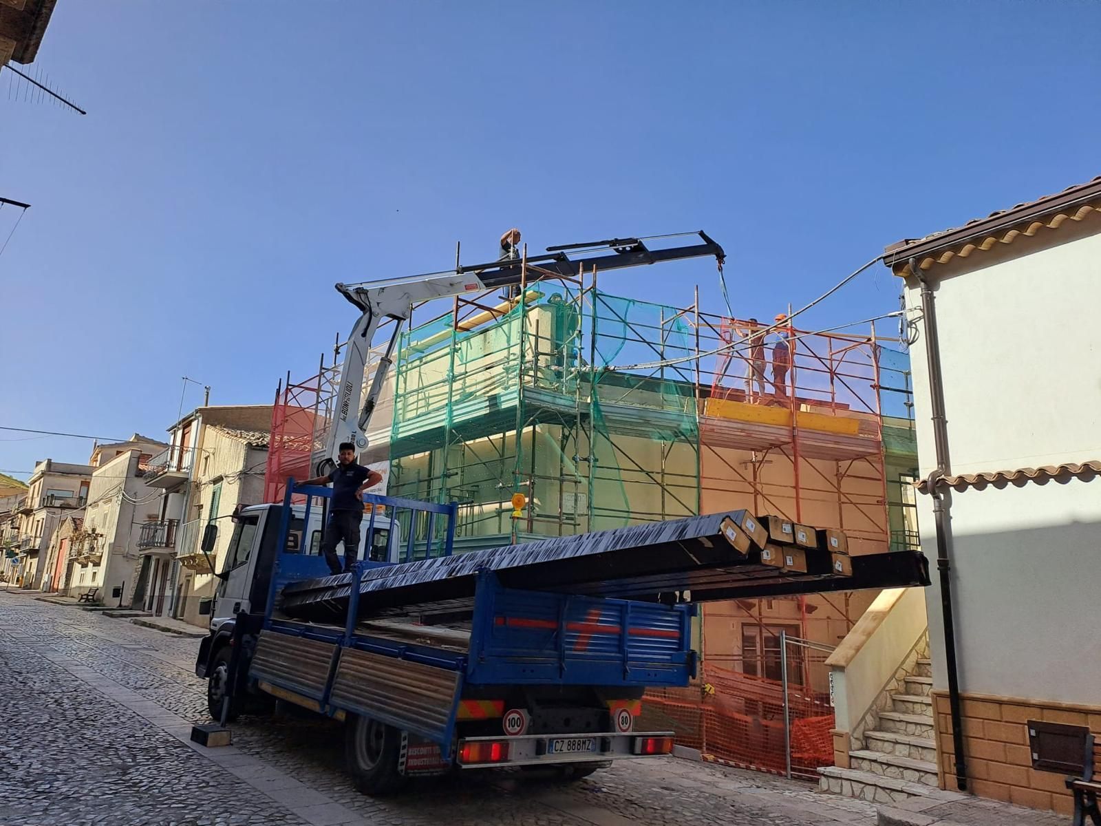 Camion con gru che trasporta grandi casse colorate su una strada acciottolata di una città, uomo in piedi sul pianale.