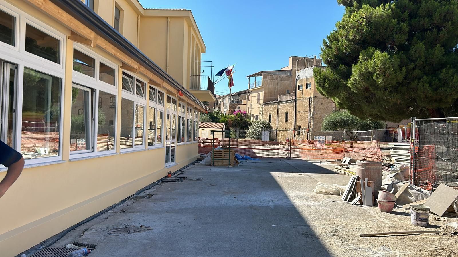 Vista esterna di un edificio in costruzione con alberi e detriti. Una bandiera è visibile in lontananza.