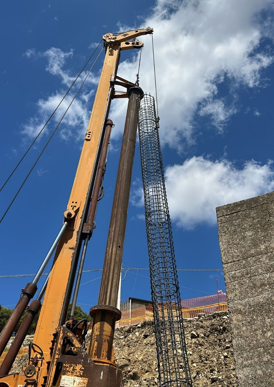 Una gru da costruzione arancione perfora vicino a un edificio in pietra sotto un cielo azzurro con nuvole.