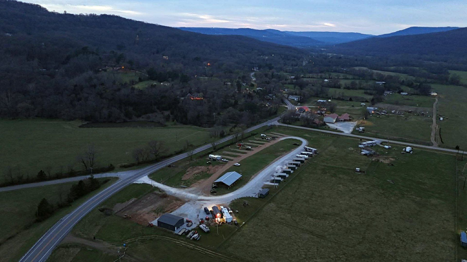An aerial view of a small RV campground at twilight, located in a valley between rolling, tree-covered hills.