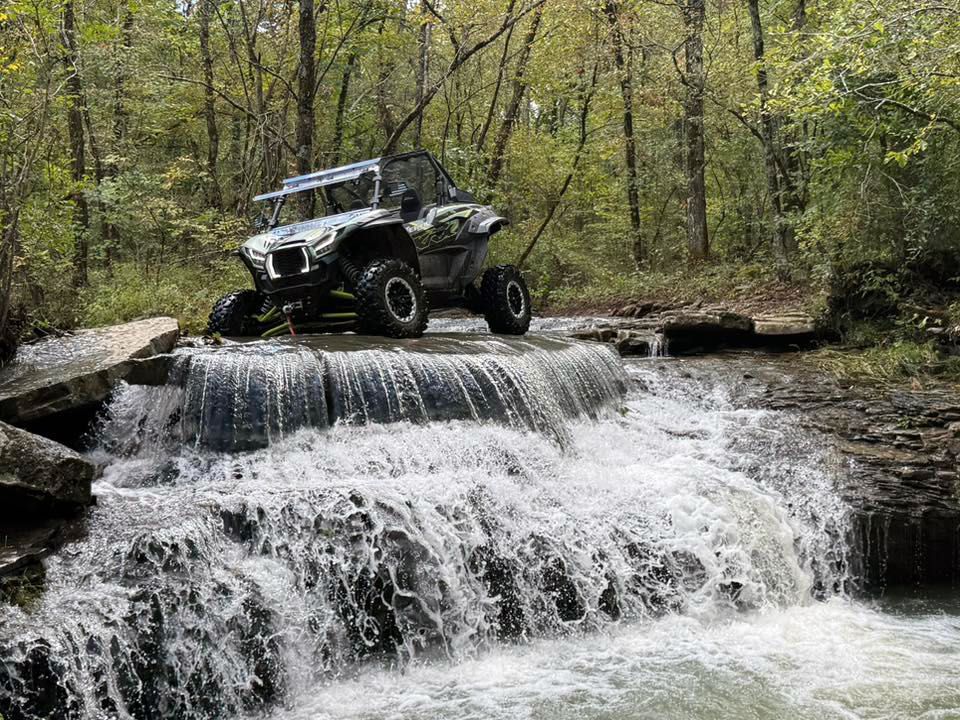 A camouflage side-by-side off-road vehicle driving across the top of a small forest waterfall.