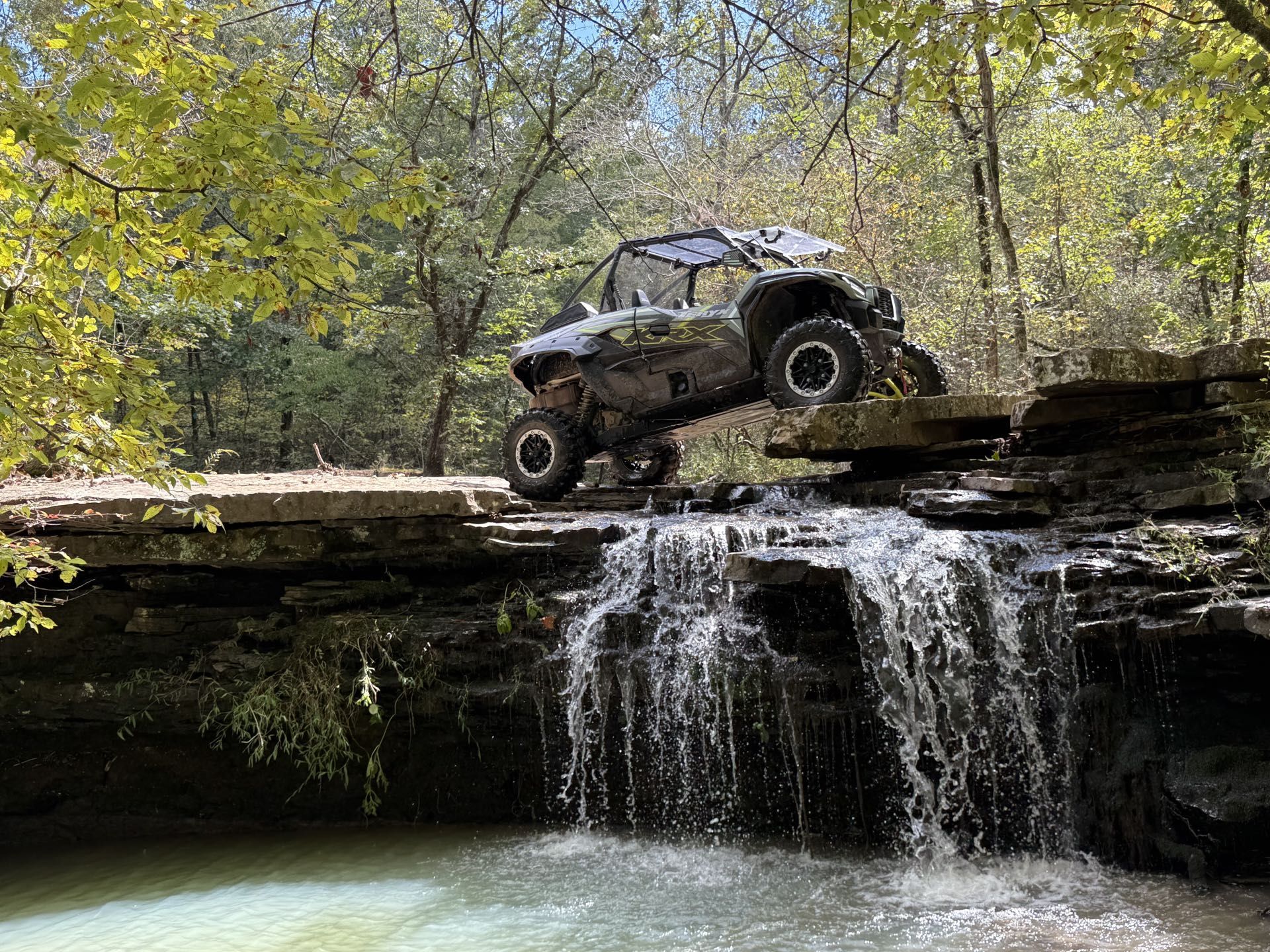An off-road vehicle perched precariously on a rocky ledge above a small waterfall in a lush, wooded area.