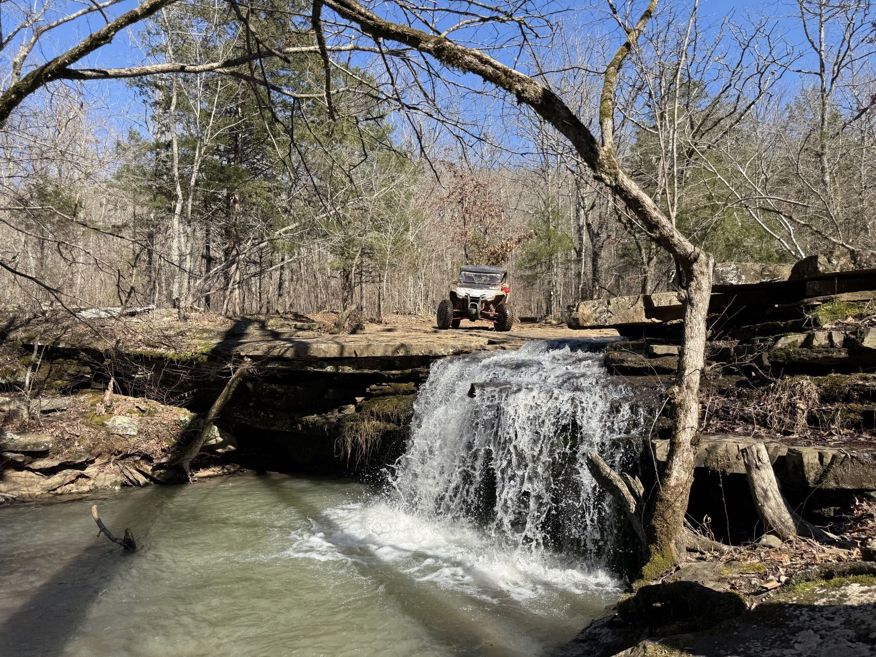 A small waterfall flows over a rocky ledge in a forest with an off-road vehicle parked on the path above.