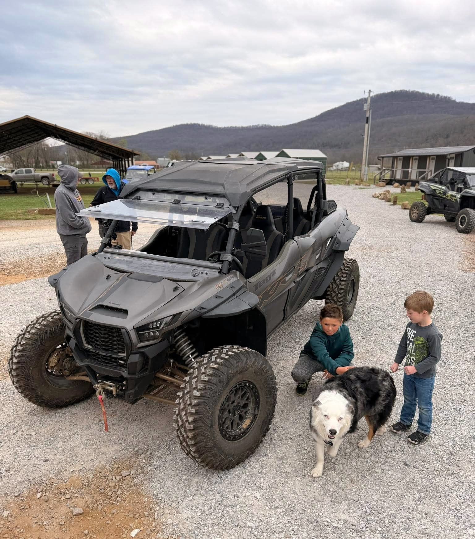 A dark gray UTV parked on a gravel lot with two people standing behind it and two children petting a dog in the front.