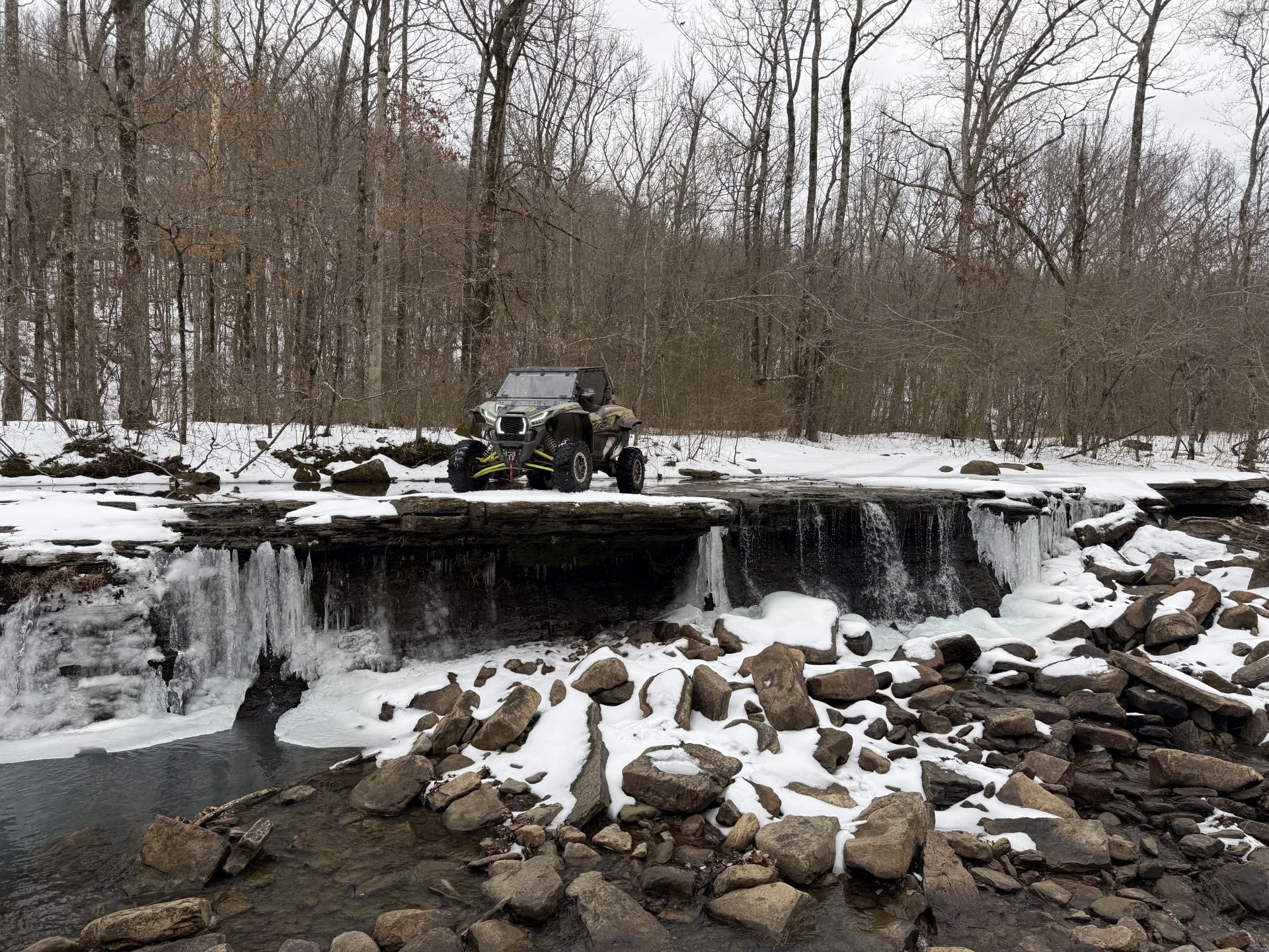 A camouflage utility vehicle parked on a frozen rock ledge above a small waterfall in a snowy, wooded landscape.