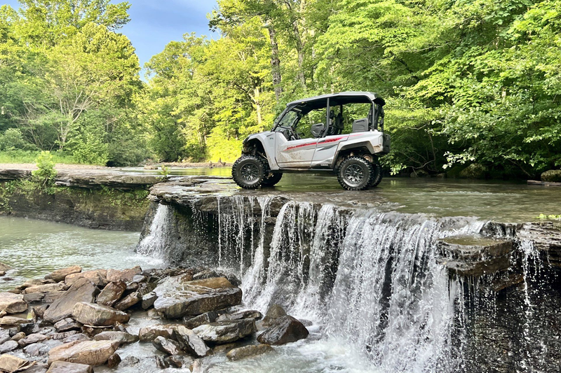 A camouflaged UTV parked on a rocky, snow-covered ledge above a partially frozen waterfall in a winter forest.