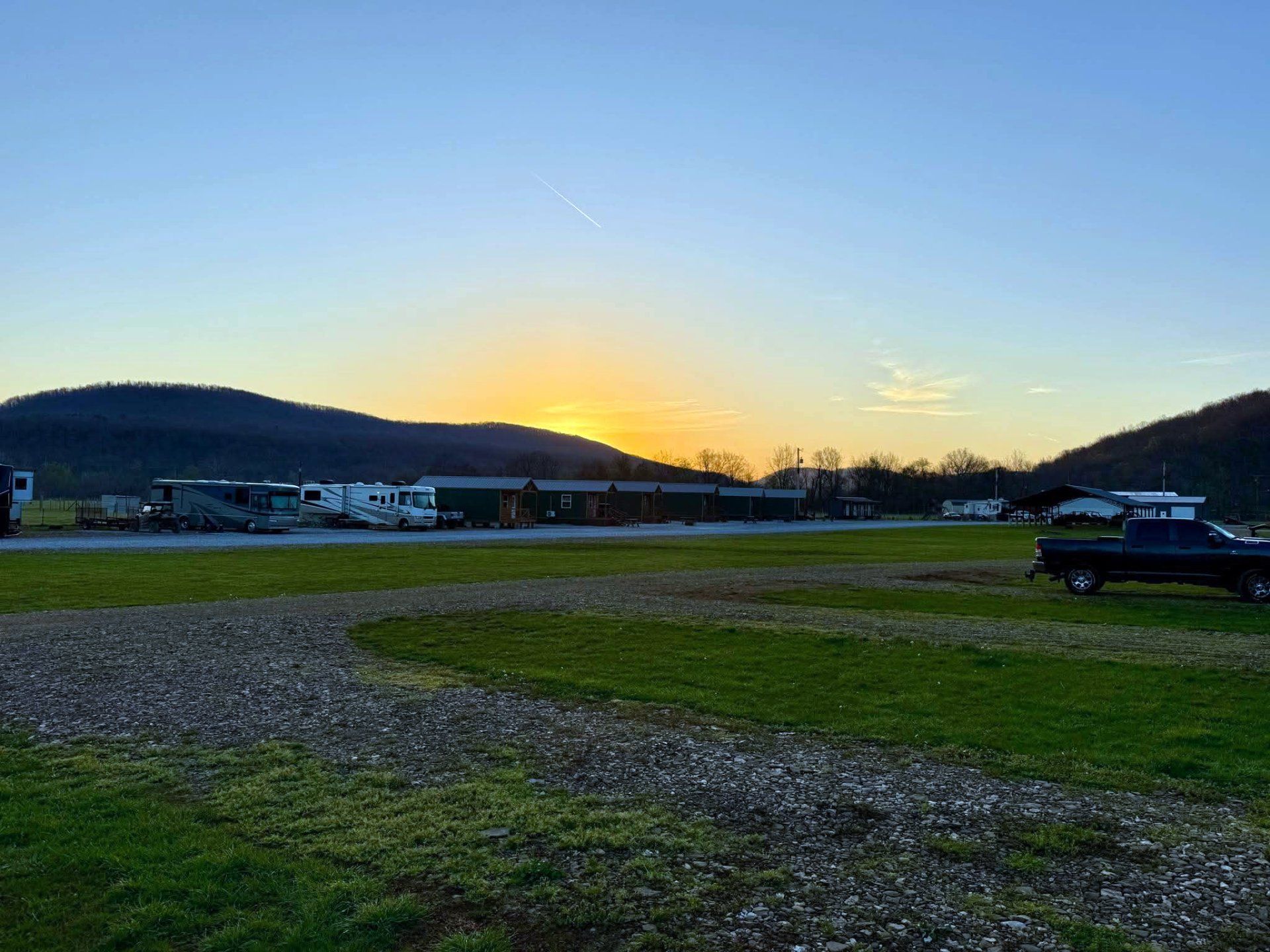 A sunset glows behind dark hills over a grassy field with parked RVs and a truck in the foreground.