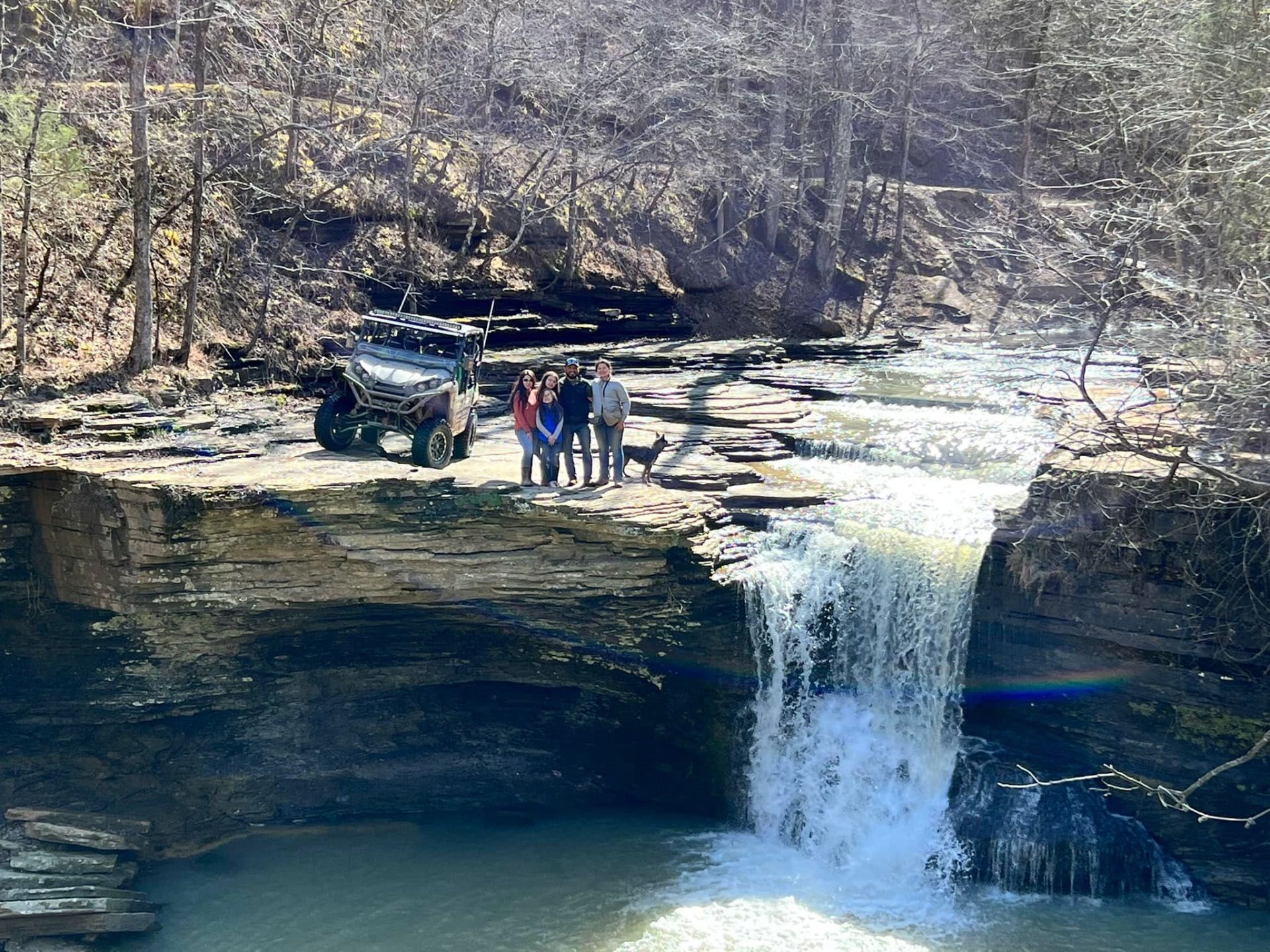A group of people and an ATV stand at the top of a rocky cliff overlooking a waterfall in a wooded area.