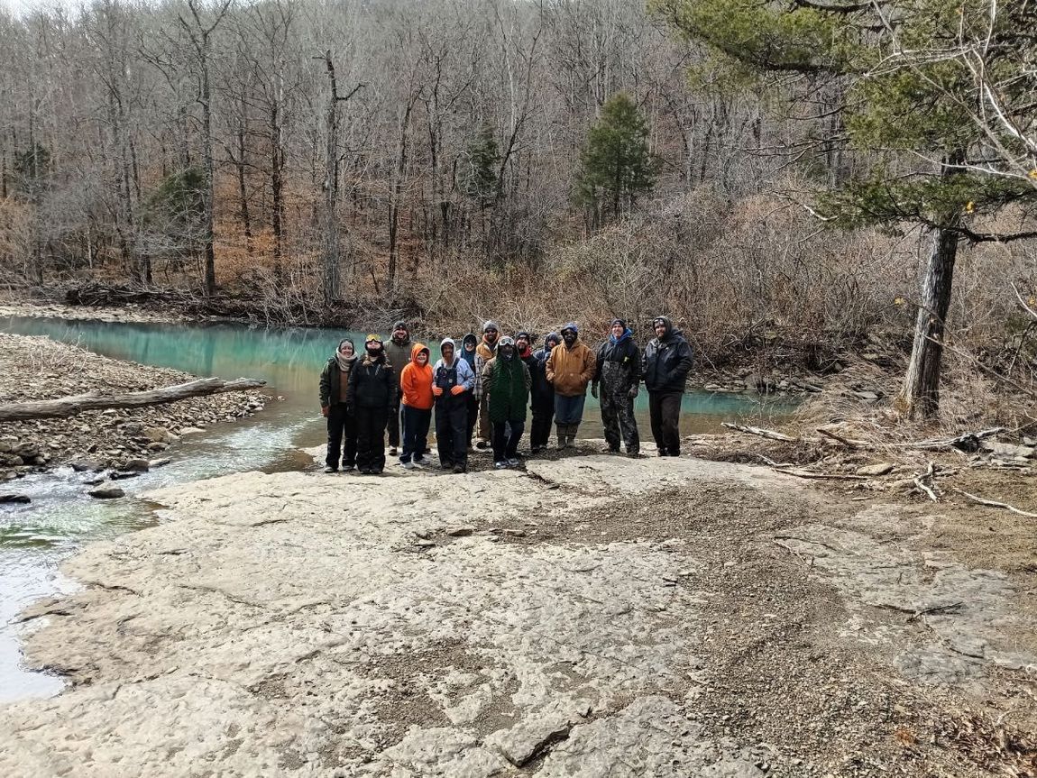 A group of people stands on a rocky riverbank in front of a turquoise stream and a forest during autumn.