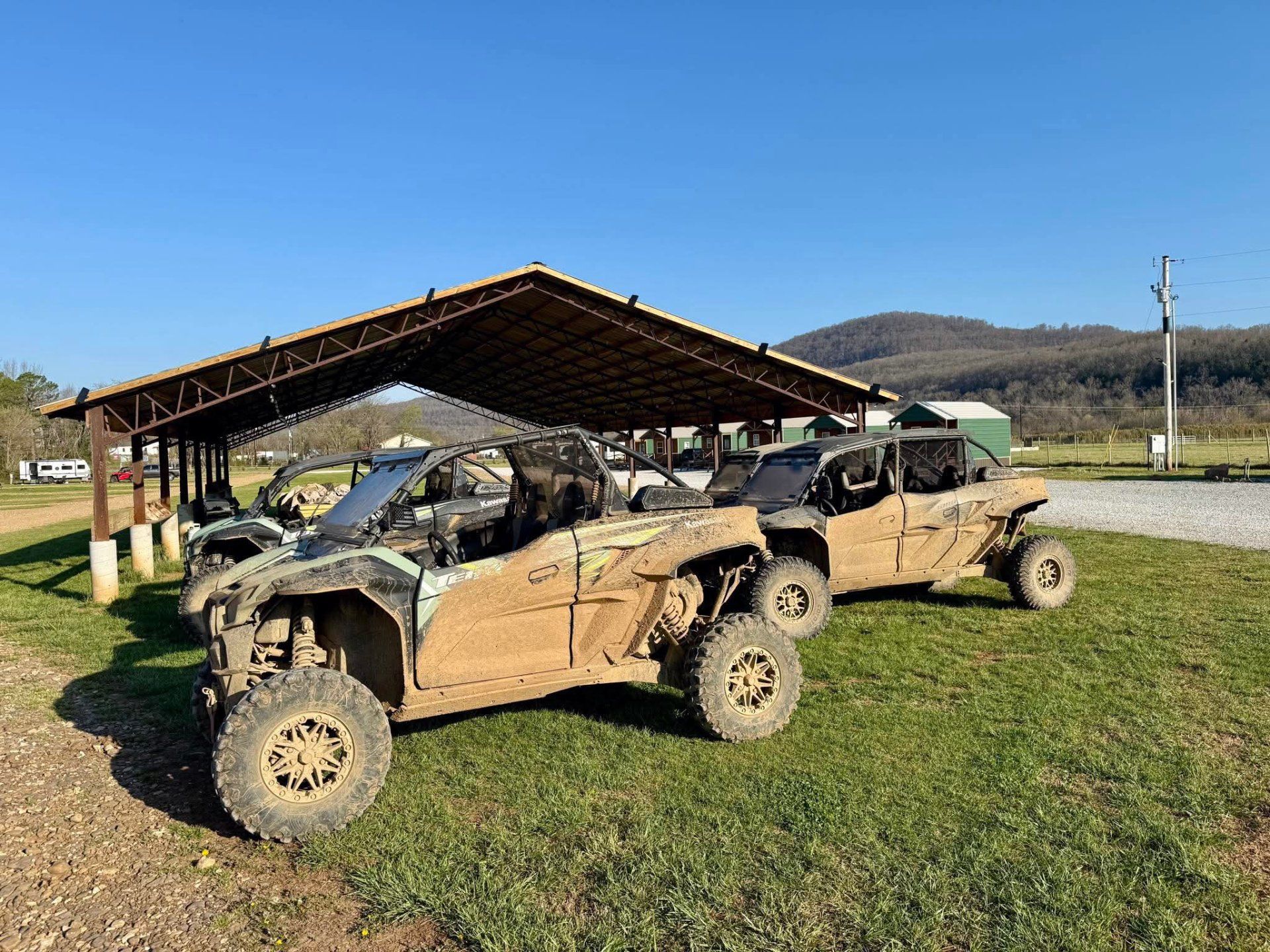 Two dusty off-road utility vehicles parked under a wooden shelter on a sunny day with hills in the background.