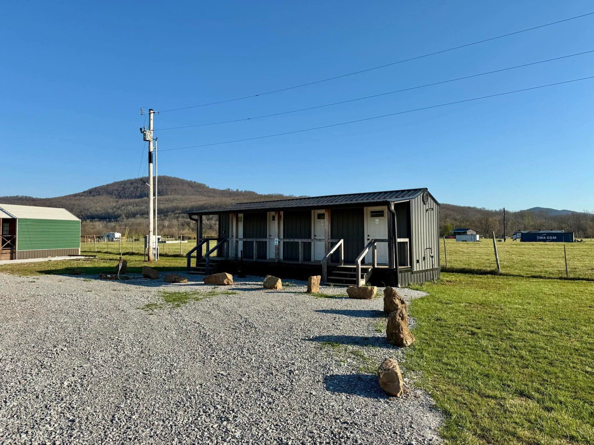 A long, gray cabin with multiple doors and a front porch sits on a gravel lot before a grassy field and distant hills.