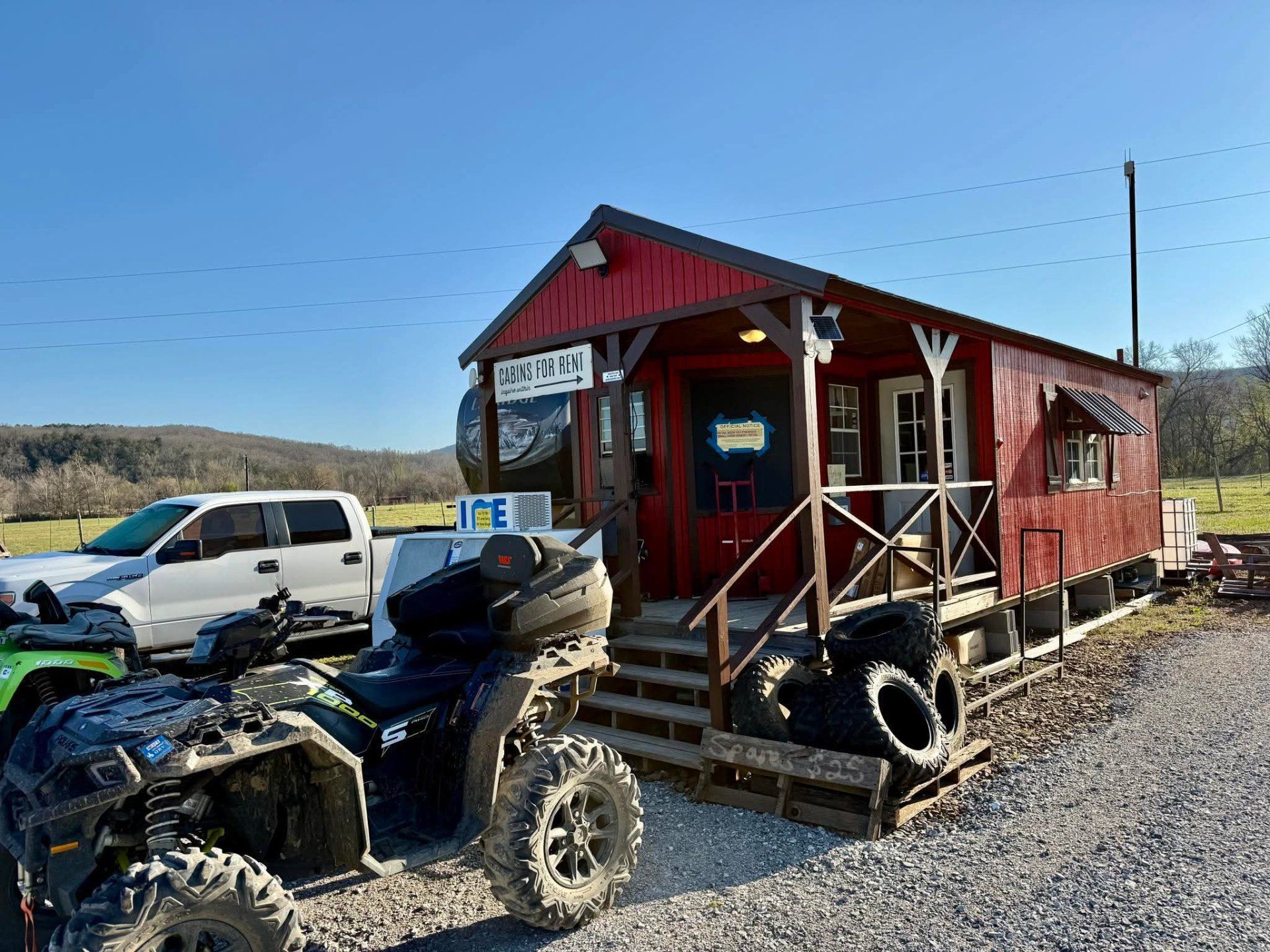 A red, wood-sided building with a porch and steps sits on a gravel lot next to an ATV and a white pickup truck.