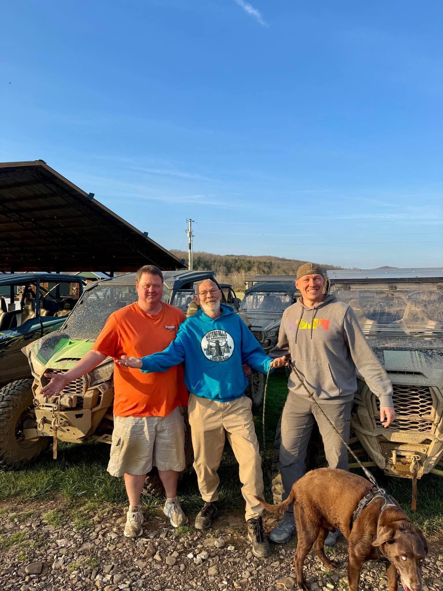 Three people and a dog stand outdoors near two off-road vehicles under a wooden structure on a sunny day.