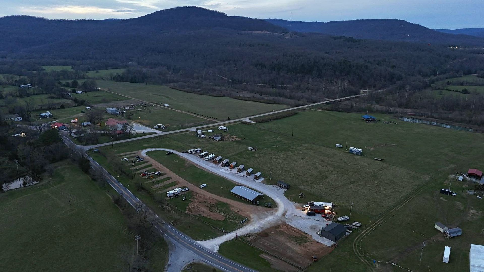 Aerial view of a rural construction site beside fields and forested hills at dusk