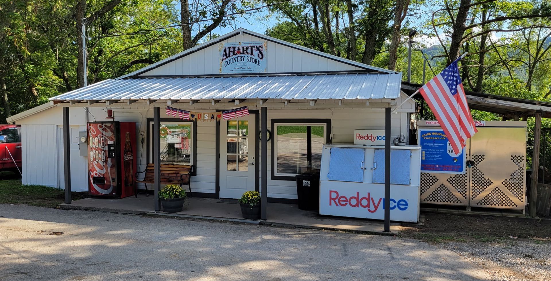 A small, white storefront with a front porch, American flag, and a Reddy Ice cooler, located outdoors on a gravel lot.