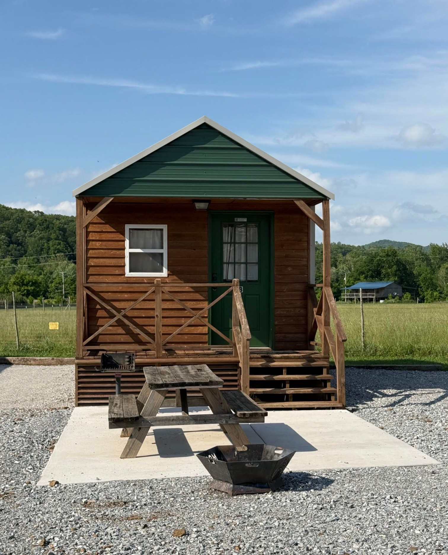 Two small, wooden camping cabins with green roofs and porches sit on a gravel ground under a clear blue sky.