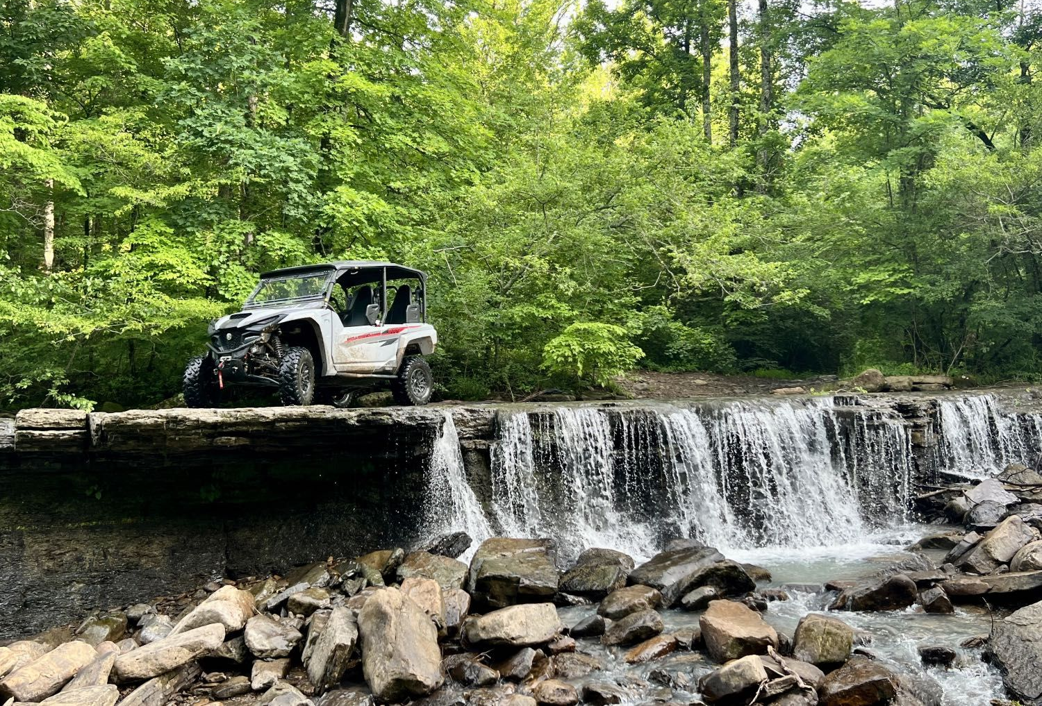 A camouflage UTV sits on a frozen, snow-covered rock ledge overlooking a small waterfall in a winter forest.