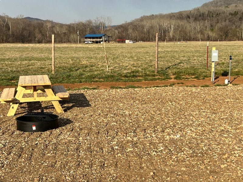A picnic table and fire ring on a gravel campsite overlooking a grassy field with a distant building and mountains.