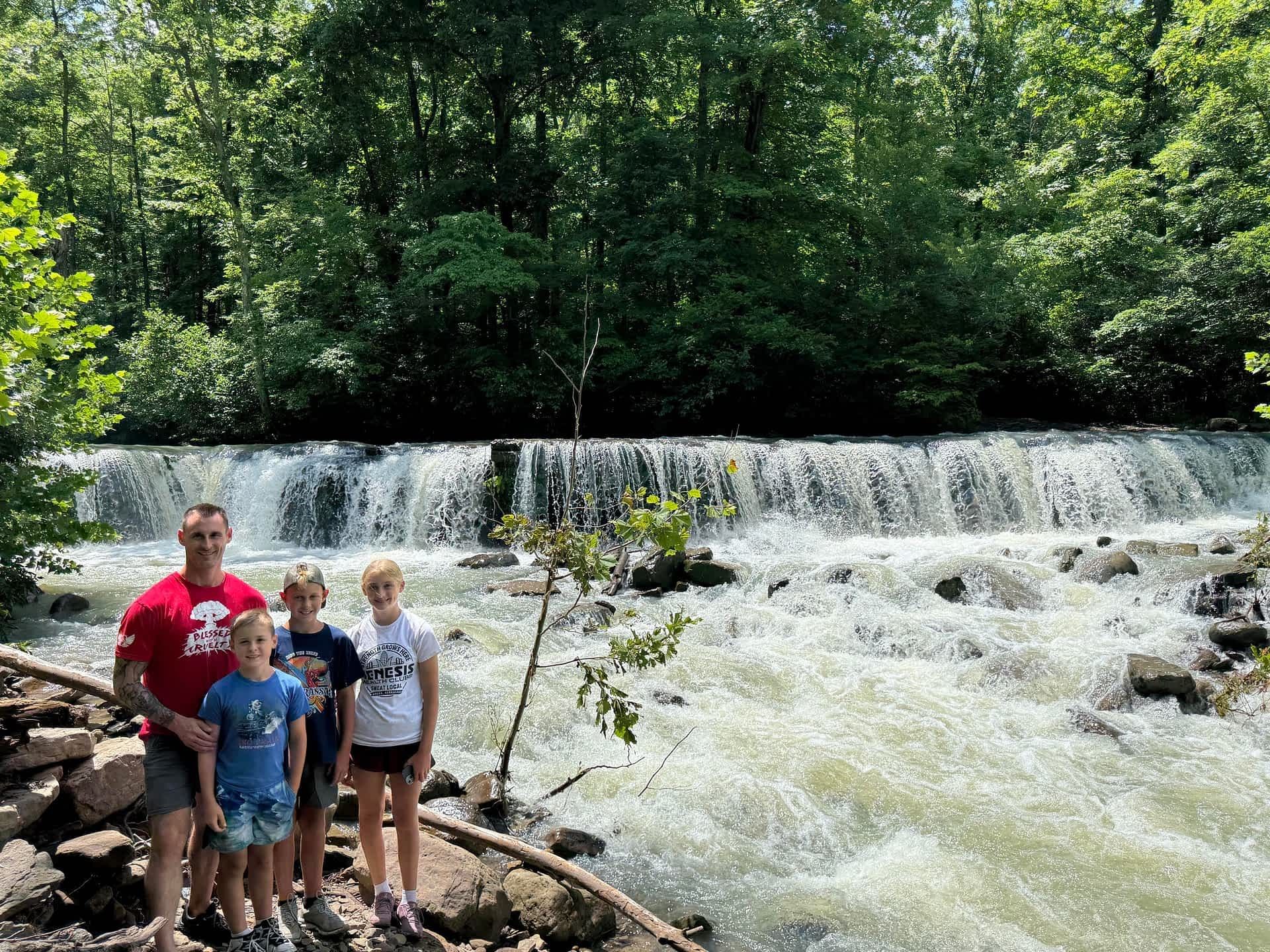 Four people posing by a foaming waterfall in a wooded park