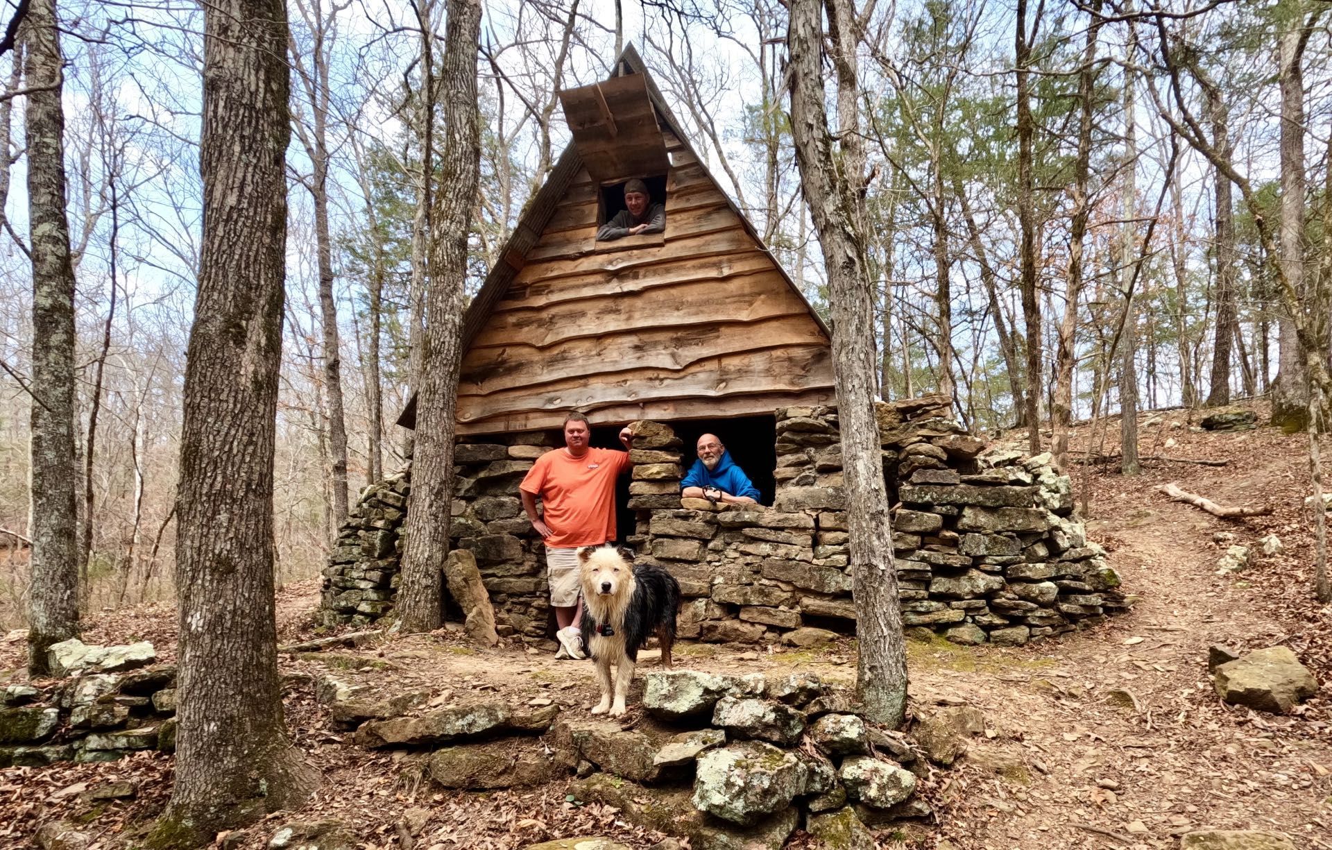 Two people and a dog stand near a rustic stone and wood cabin in a forest clearing.