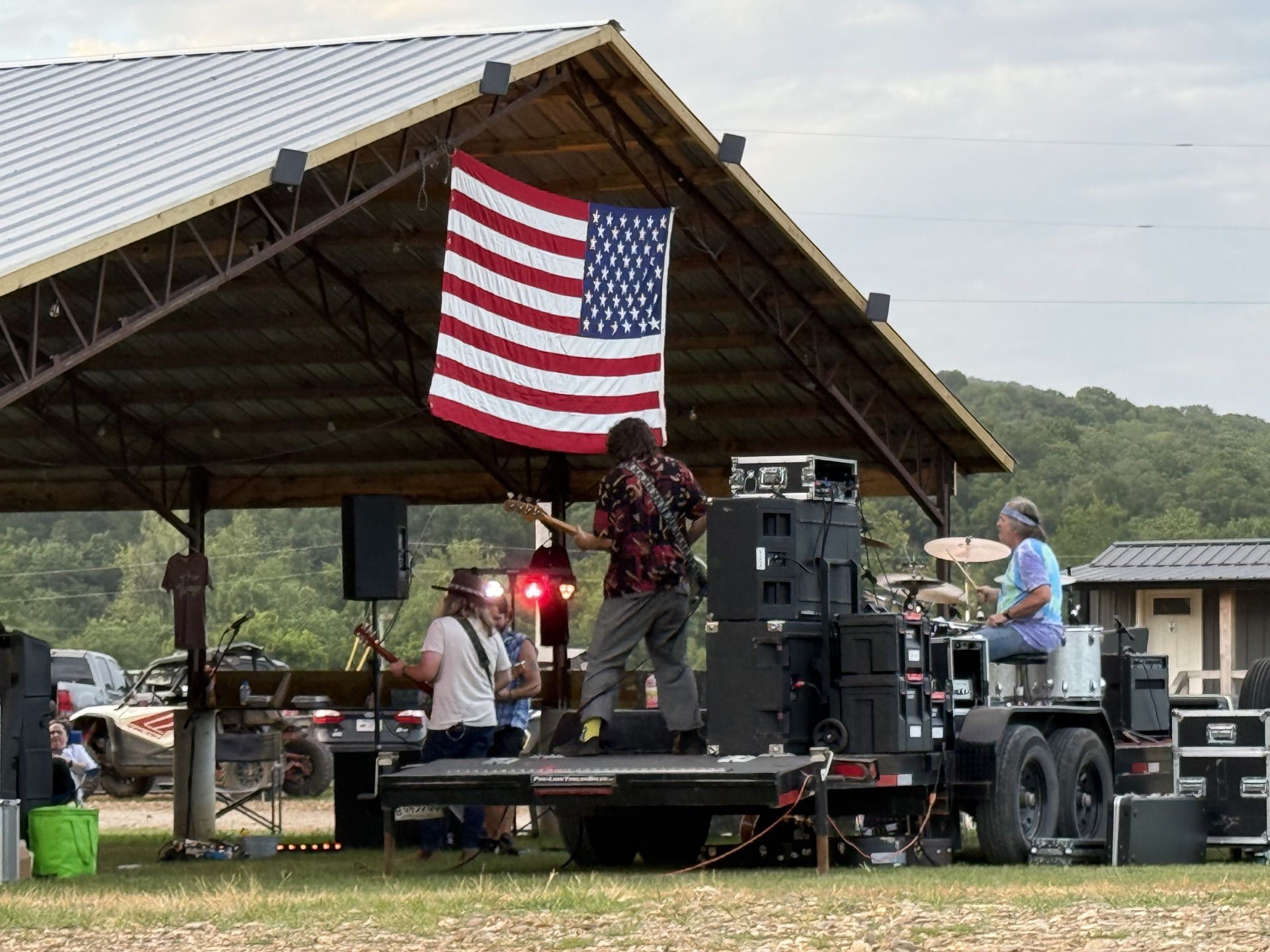 A live band performs on a trailer under a pavilion, with an American flag hanging from the roof rafters.