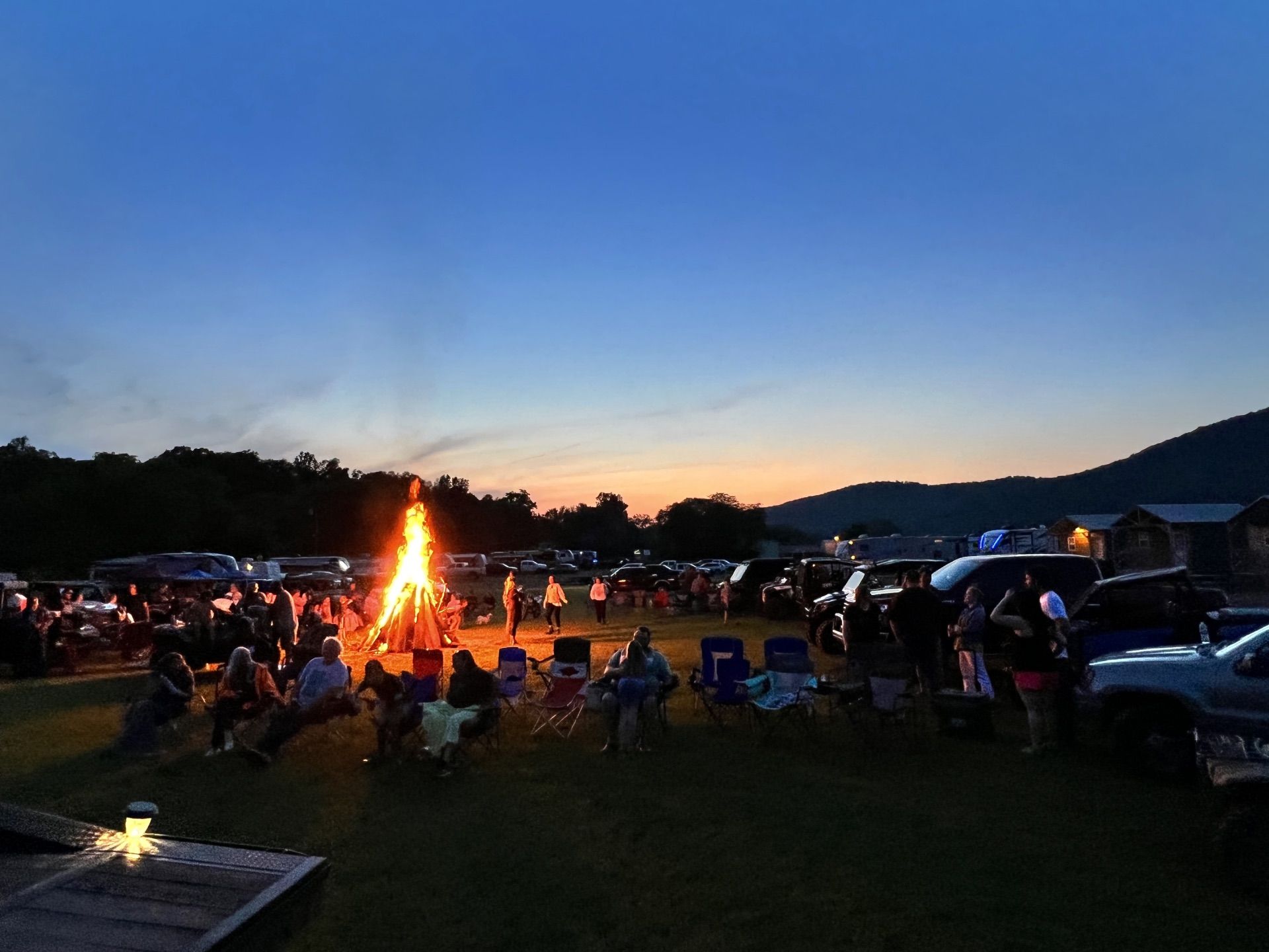 A large campfire burns brightly at twilight in a grassy field with people sitting in lawn chairs nearby and parked cars.