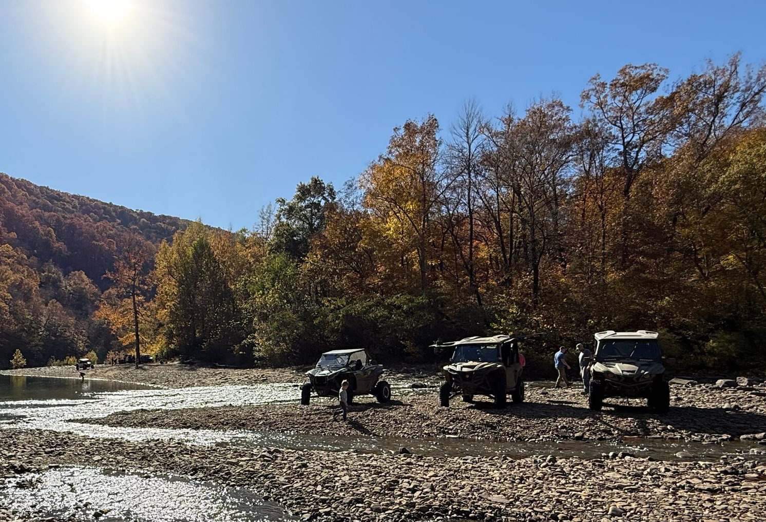 Shallow rocky river with parked off-road vehicles and autumn trees under bright sun