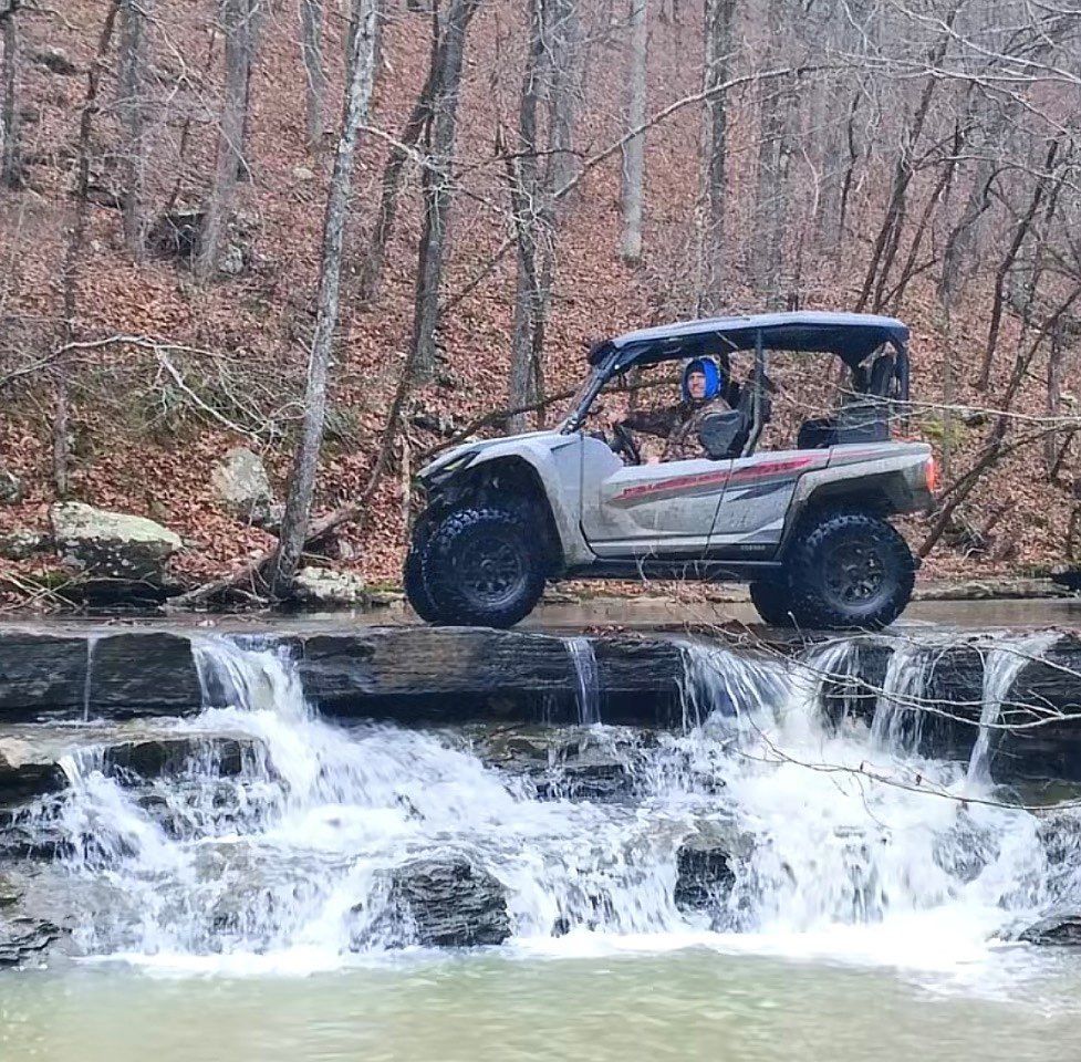 A gray UTV is parked on a flat rock ledge in the middle of a small forest waterfall.