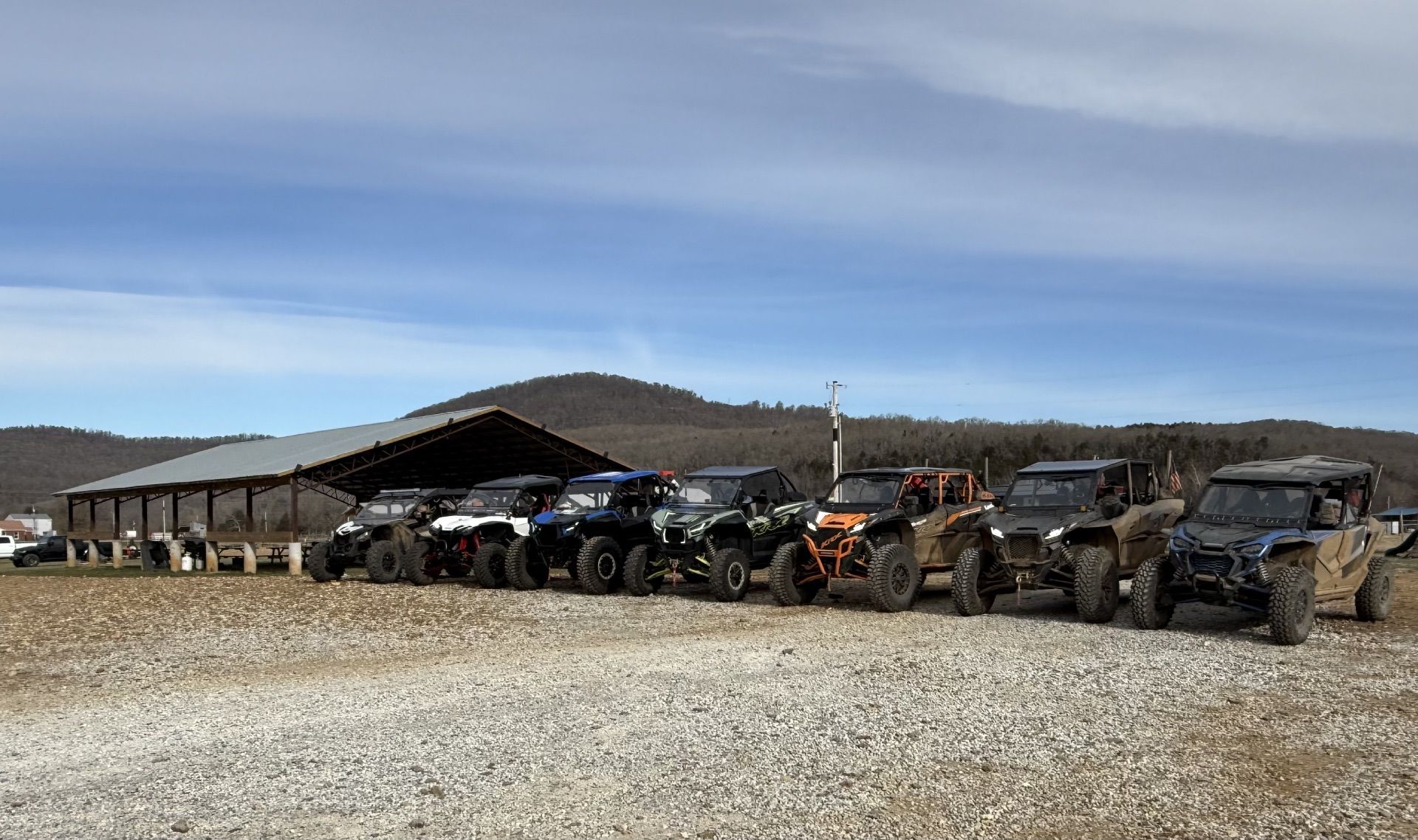A line of side-by-side UTVs parked on a gravel lot in front of a metal pavilion under a clear, blue sky.