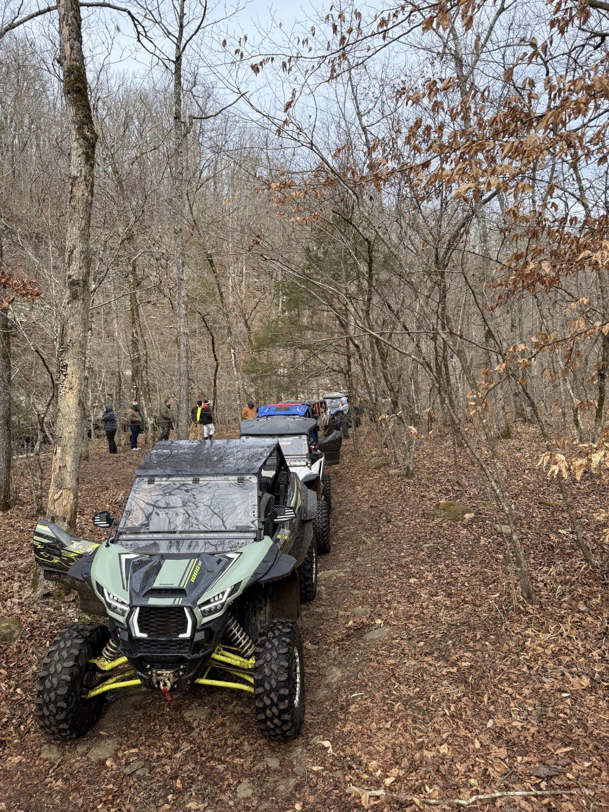 Black and green off-road UTV on a muddy wooded trail, with another vehicle behind it.