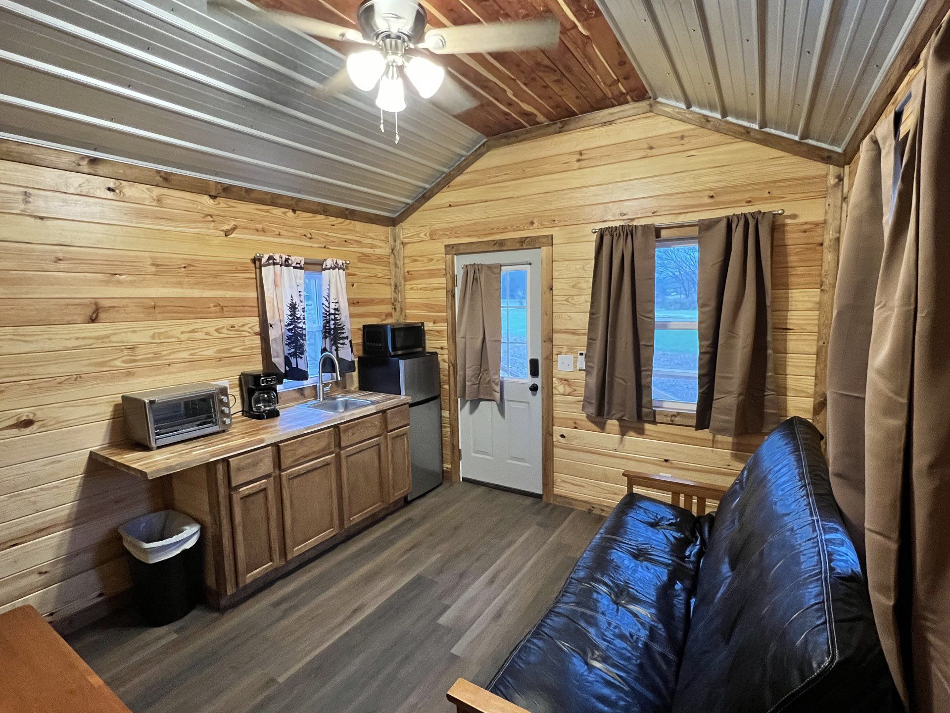 A rustic bedroom with wood-paneled walls, a ceiling fan, a window with patterned curtains, and a white bed.