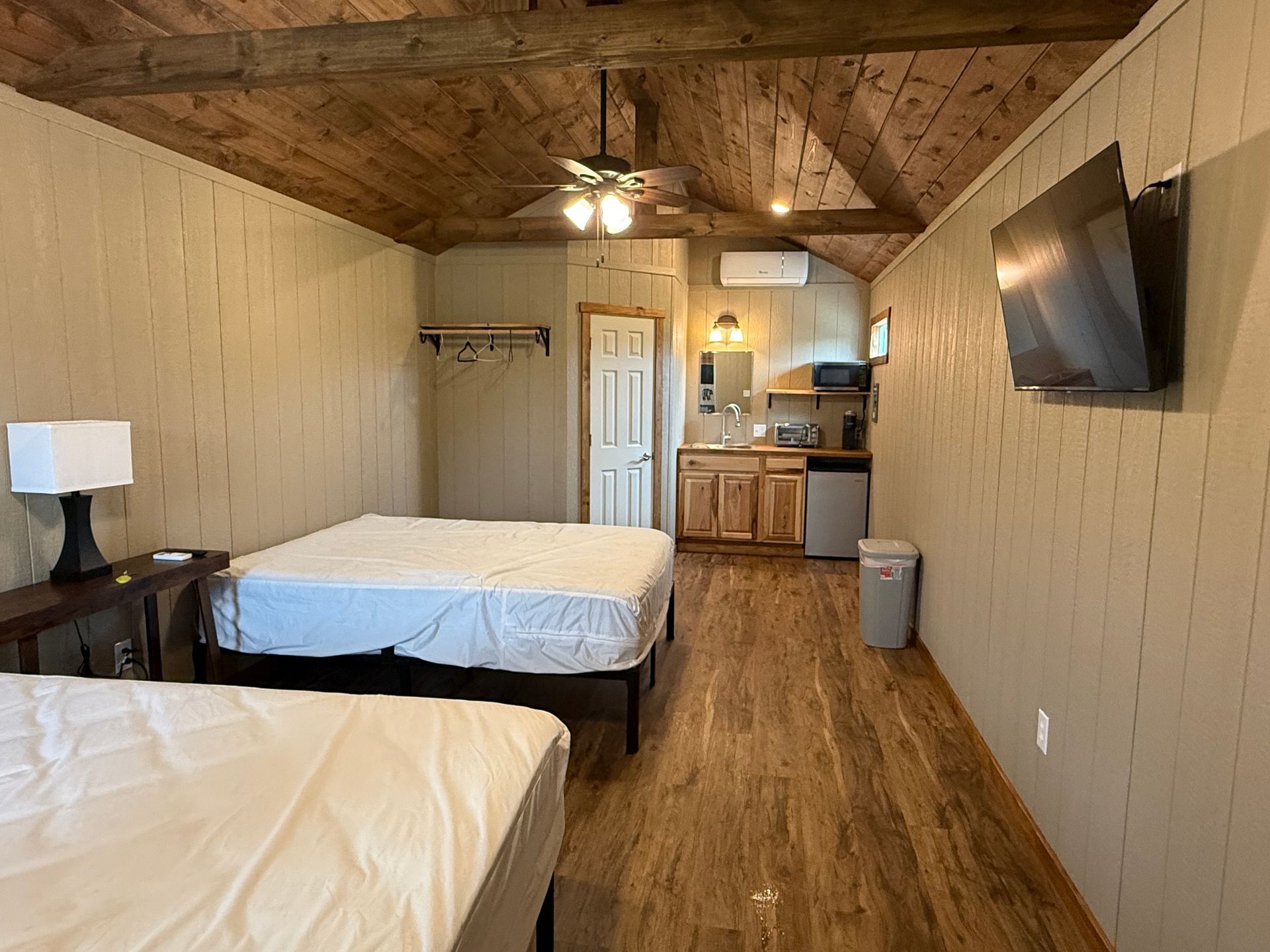 A small, rustic cabin interior with wooden walls, a bunk bed, a dining table with chairs, and a ceiling fan.