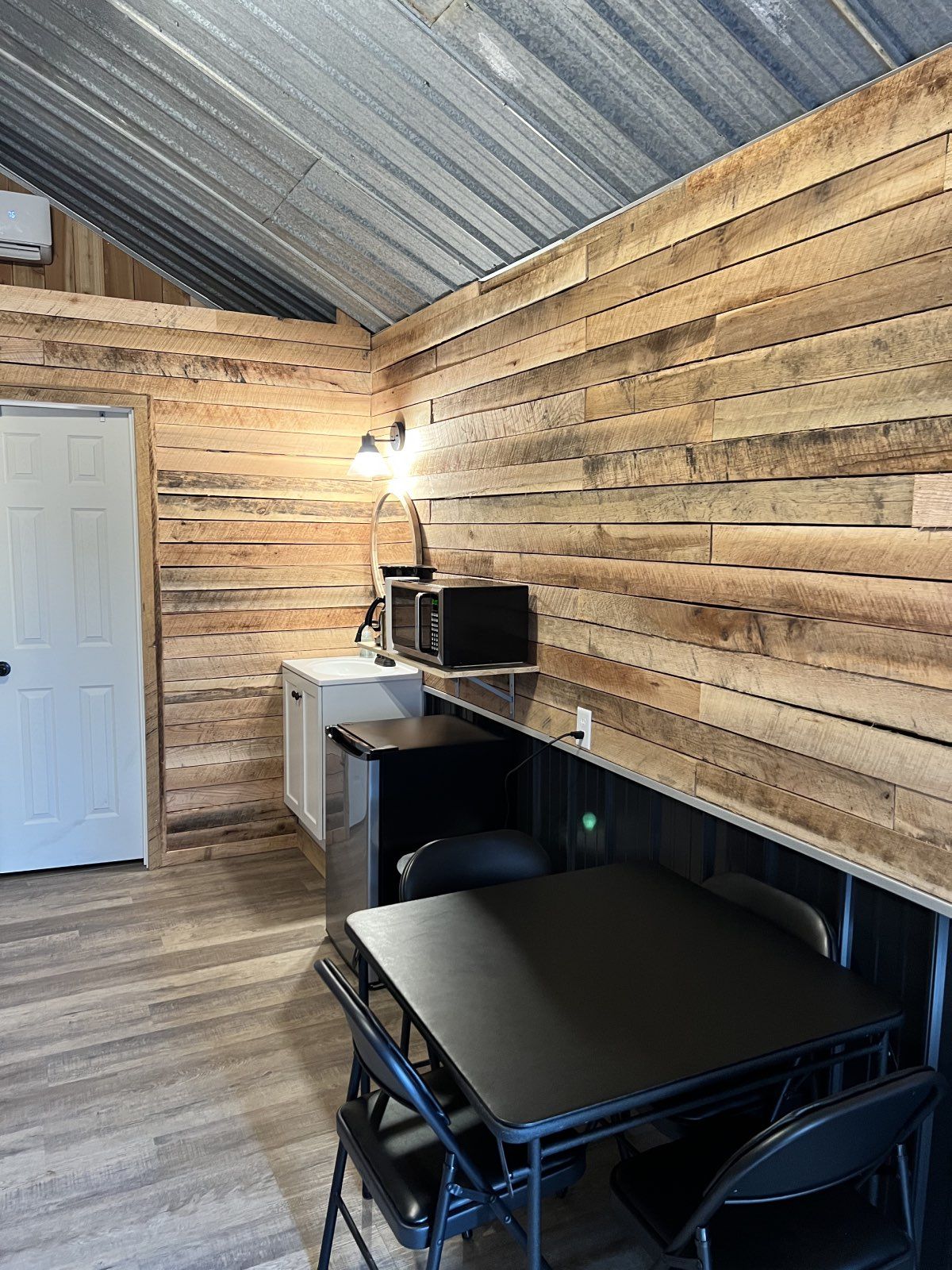 Small rustic office with wood plank walls, metal ceiling, desk, chair, and microwave by a white door.
