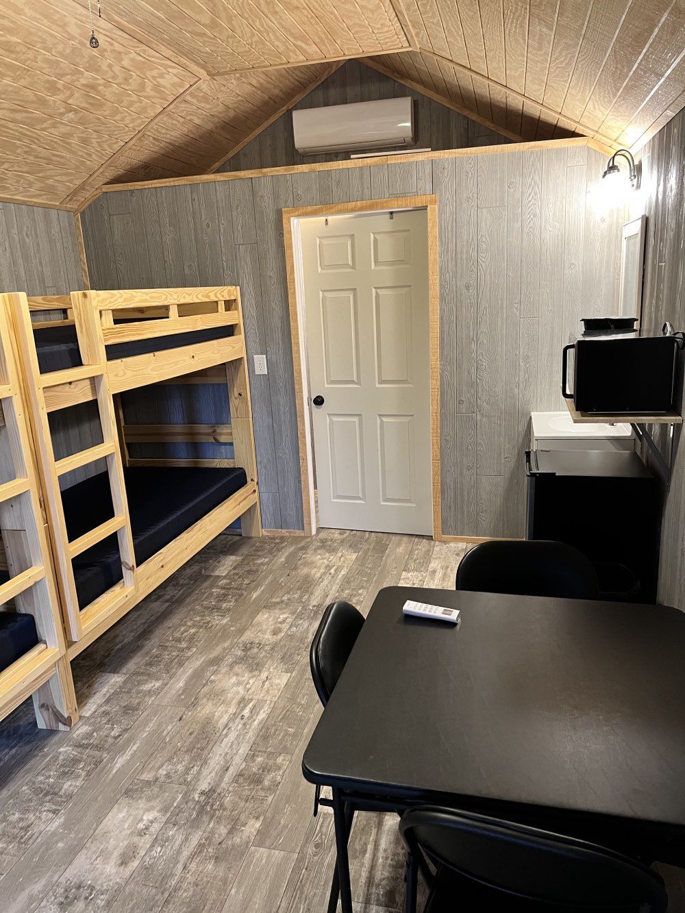 A small, rustic room with a wooden bunk bed, a black table and chairs, and a white door under a vaulted ceiling.
