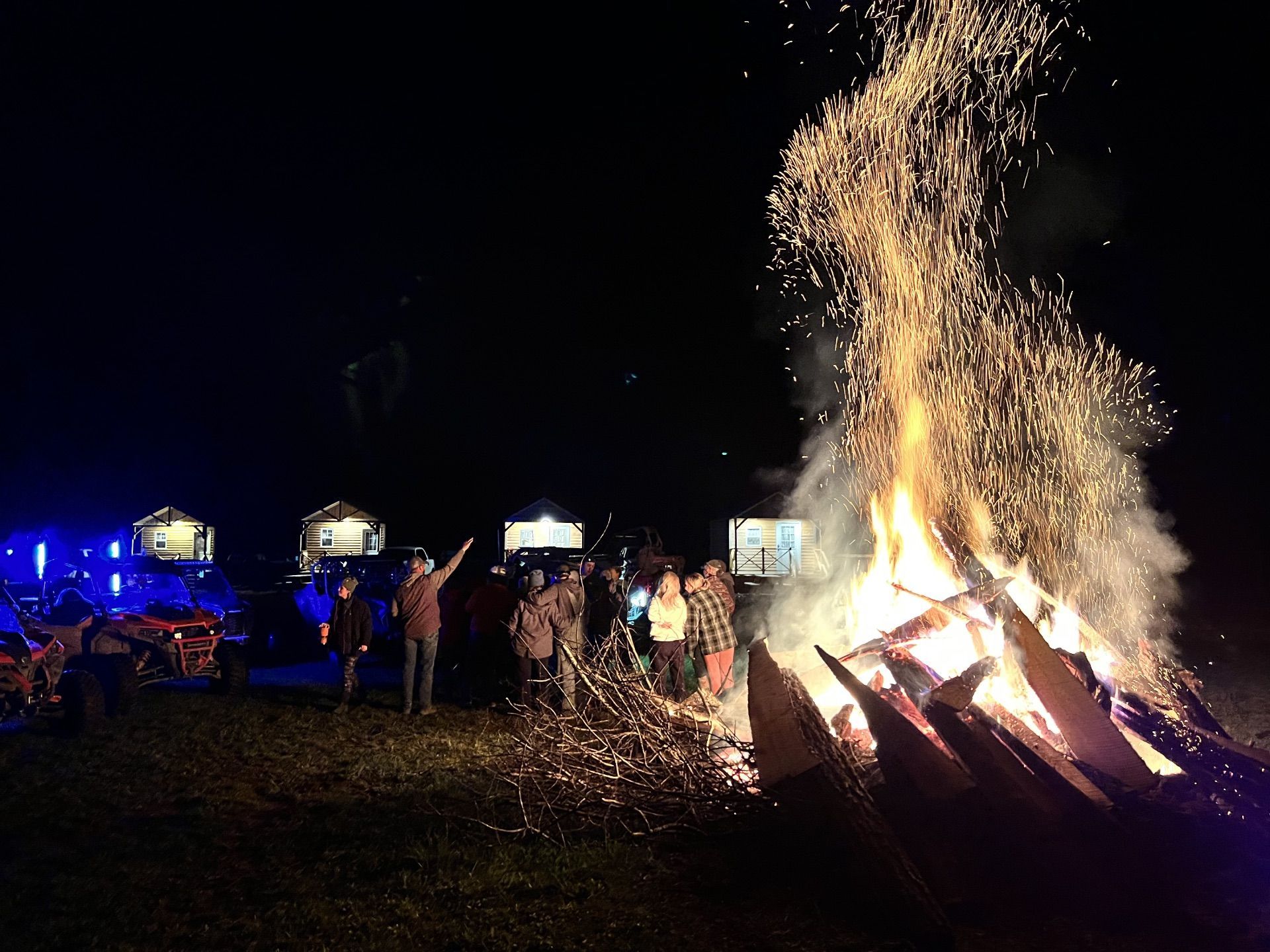 A group of people standing near a large, bright bonfire at night with sparks rising into the dark sky.