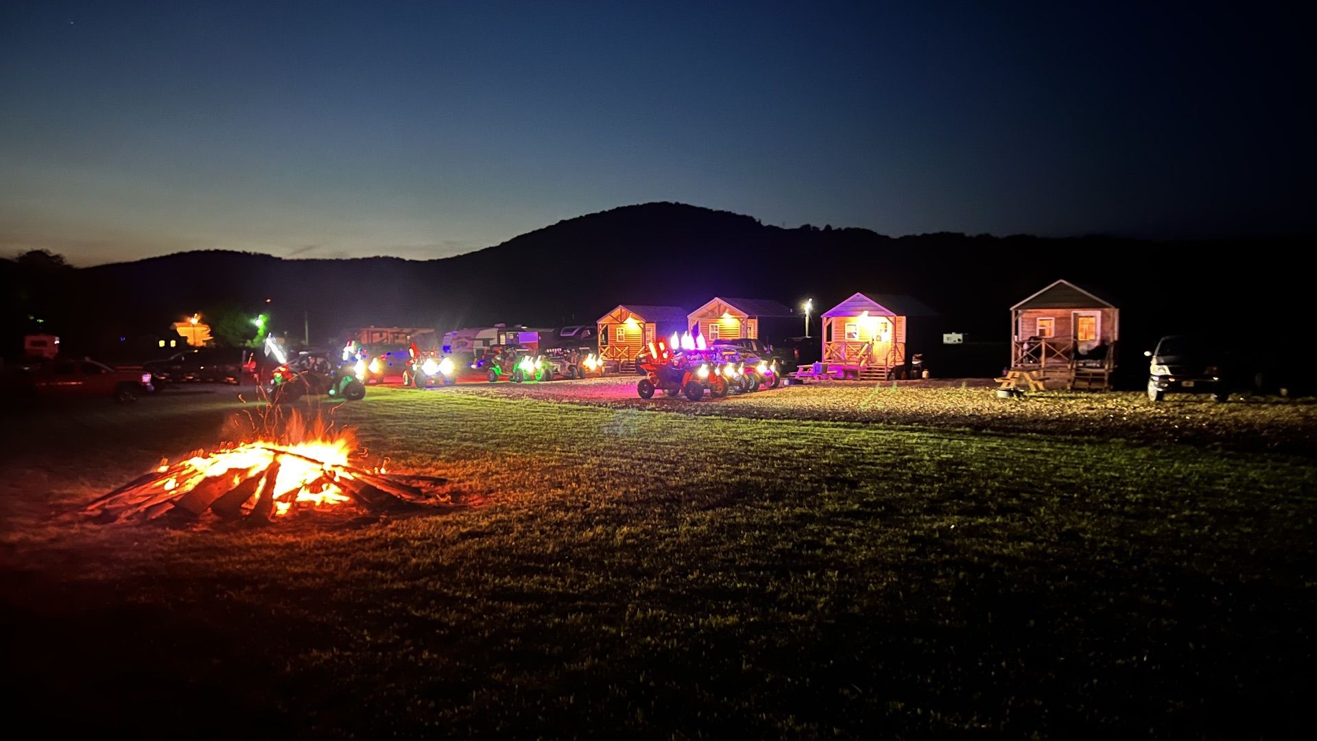 A bonfire burns in a field at night, with illuminated small cabins and vehicles parked in the background.
