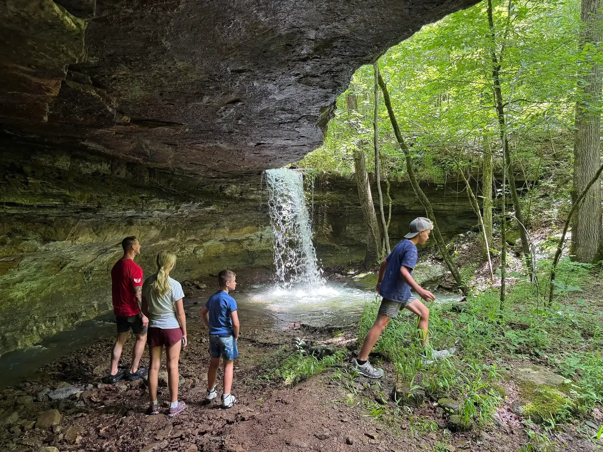 People standing near a small waterfall in a rocky forest overhang, with one person stepping forward.