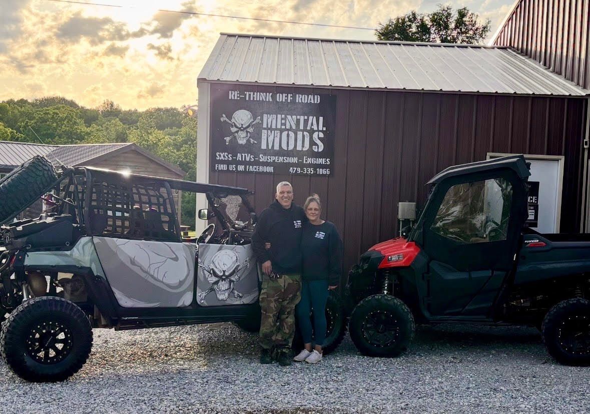 A black off-road vehicle parked on a gravel lot with two people standing behind it and two children petting a dog nearby.