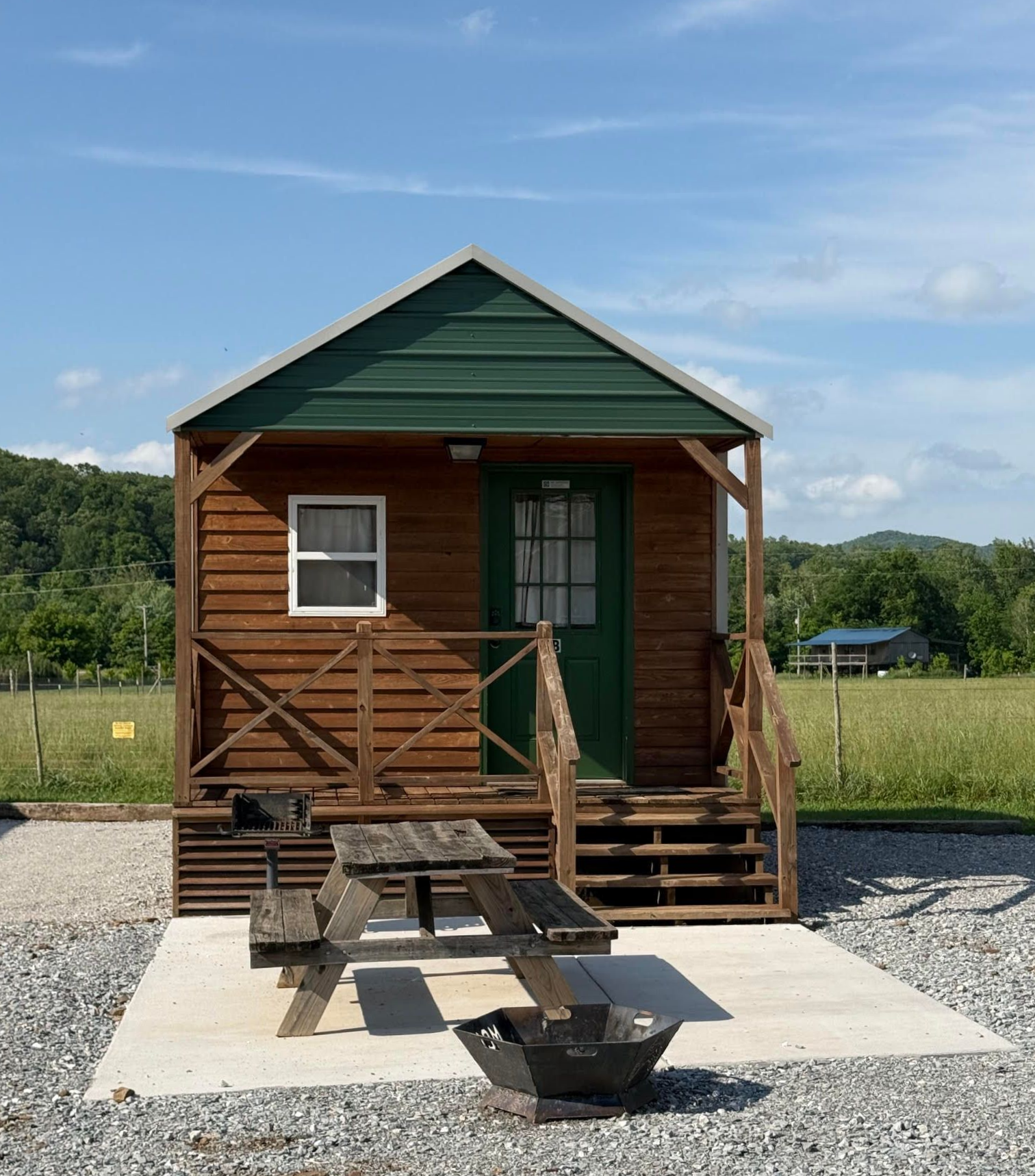 Two small, wooden camping cabins with green roofs and porches sit on a gravel ground under a clear blue sky.