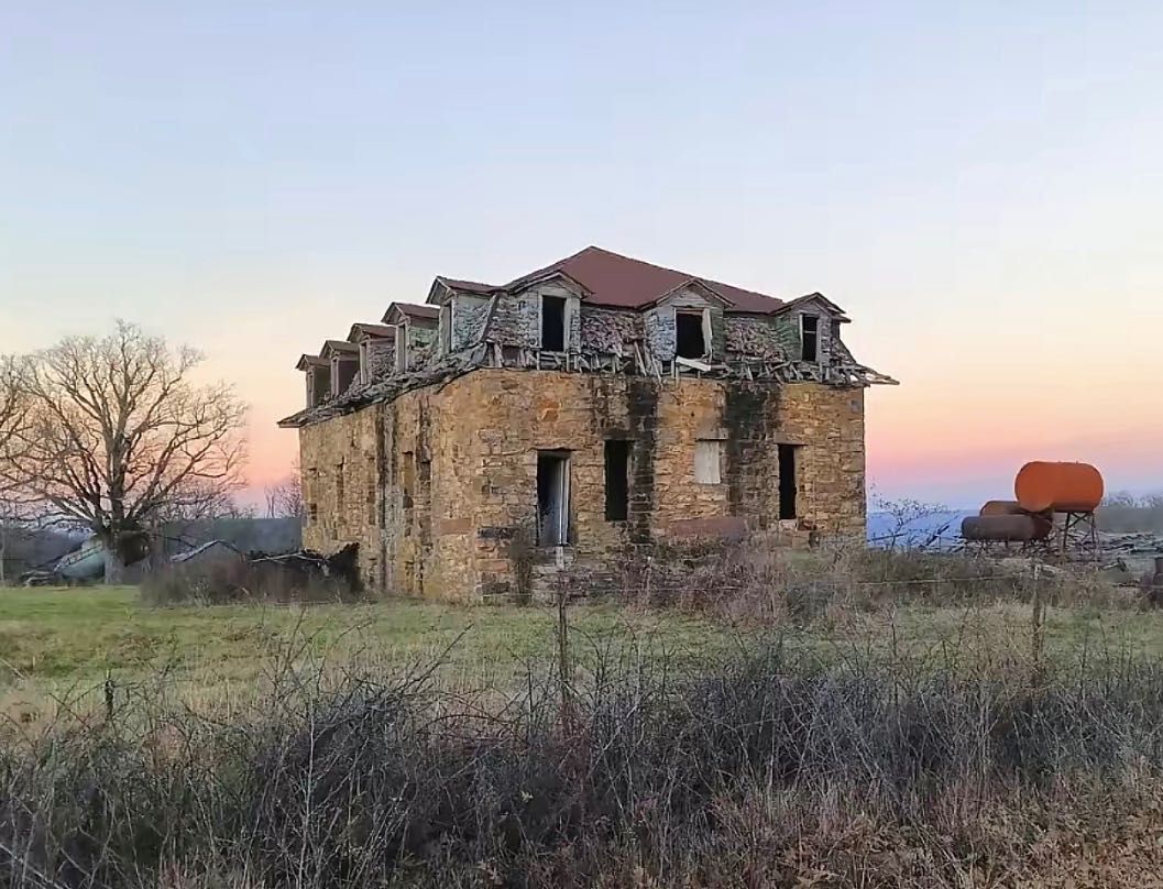 An abandoned, multi-story stone house with dormer windows stands in a field at dusk under a clear, pale sky.