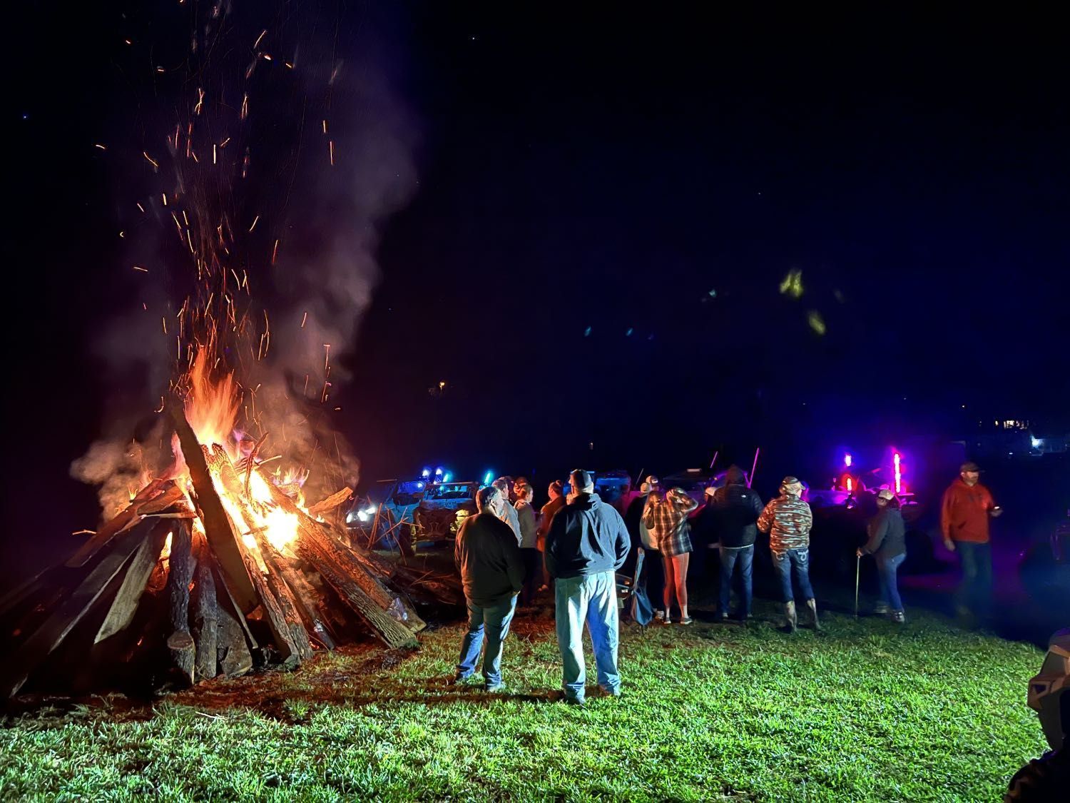 A group of people standing in a field at night, gathered around a large, bright, flaming bonfire.