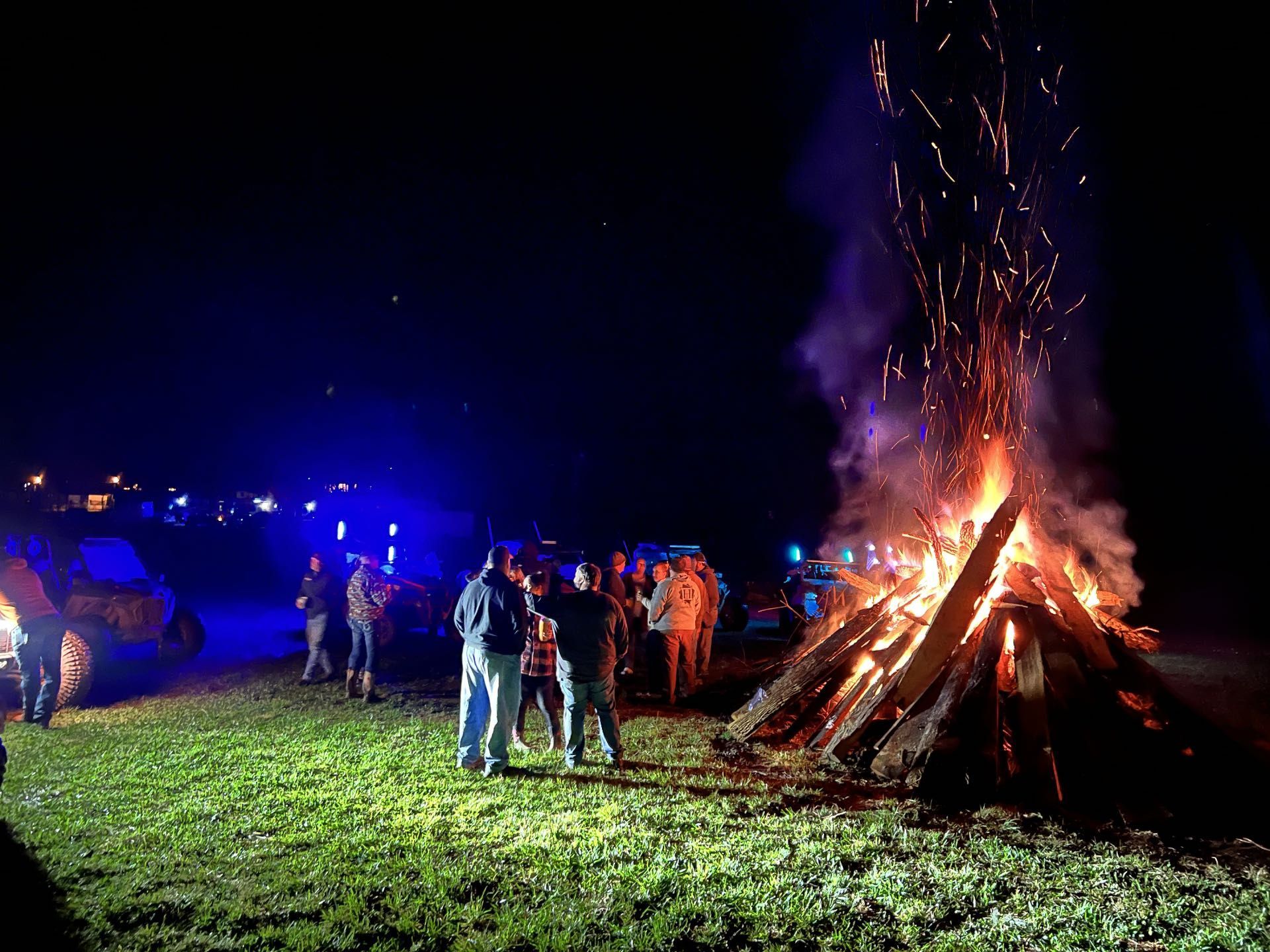 A group of people standing in a field at night near a large, bright bonfire with sparks rising into the dark sky.