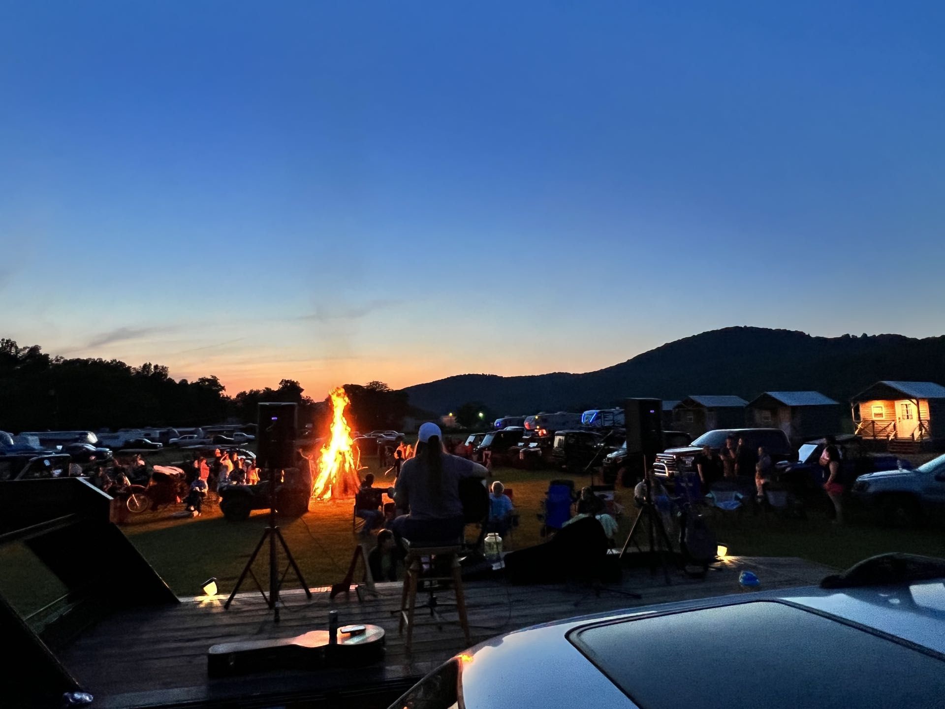 A musician performs on a stage facing an audience gathered around a campfire at dusk, with silhouettes of hills nearby.